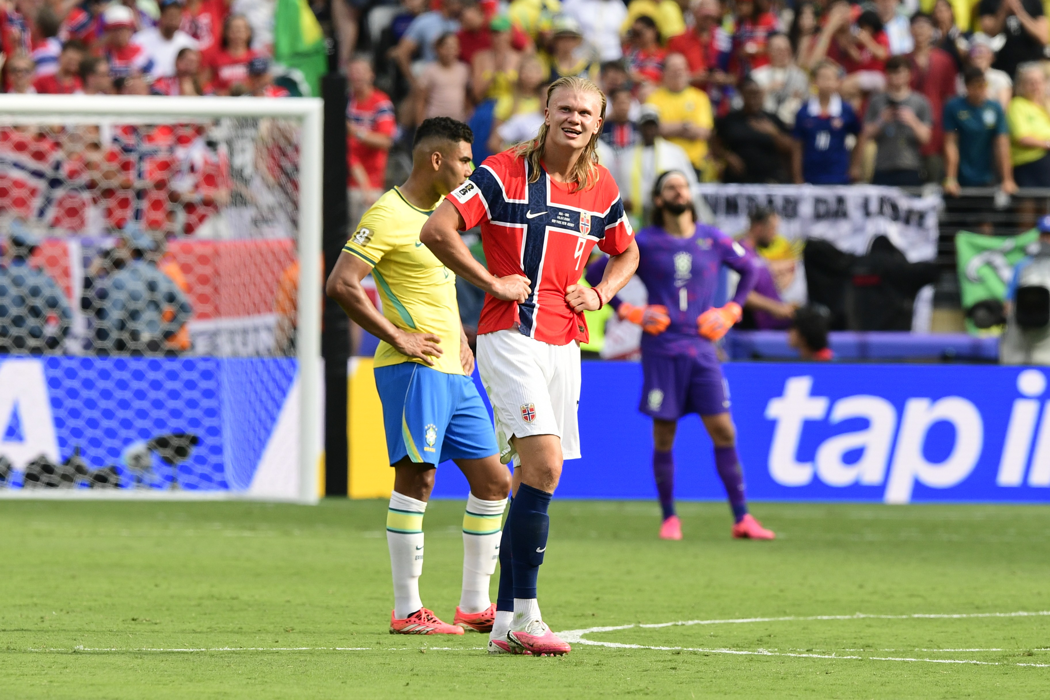 Dois jogadores de futebol, um com camisa amarela e outro com camisa vermelha, estão em campo com as mãos na cintura, olhando para frente. O jogador de camisa amarela tem cabelo escuro e barba, enquanto o de camisa vermelha tem cabelo loiro comprido. Ao fundo, um goleiro de roxo e torcedores nas arquibancadas