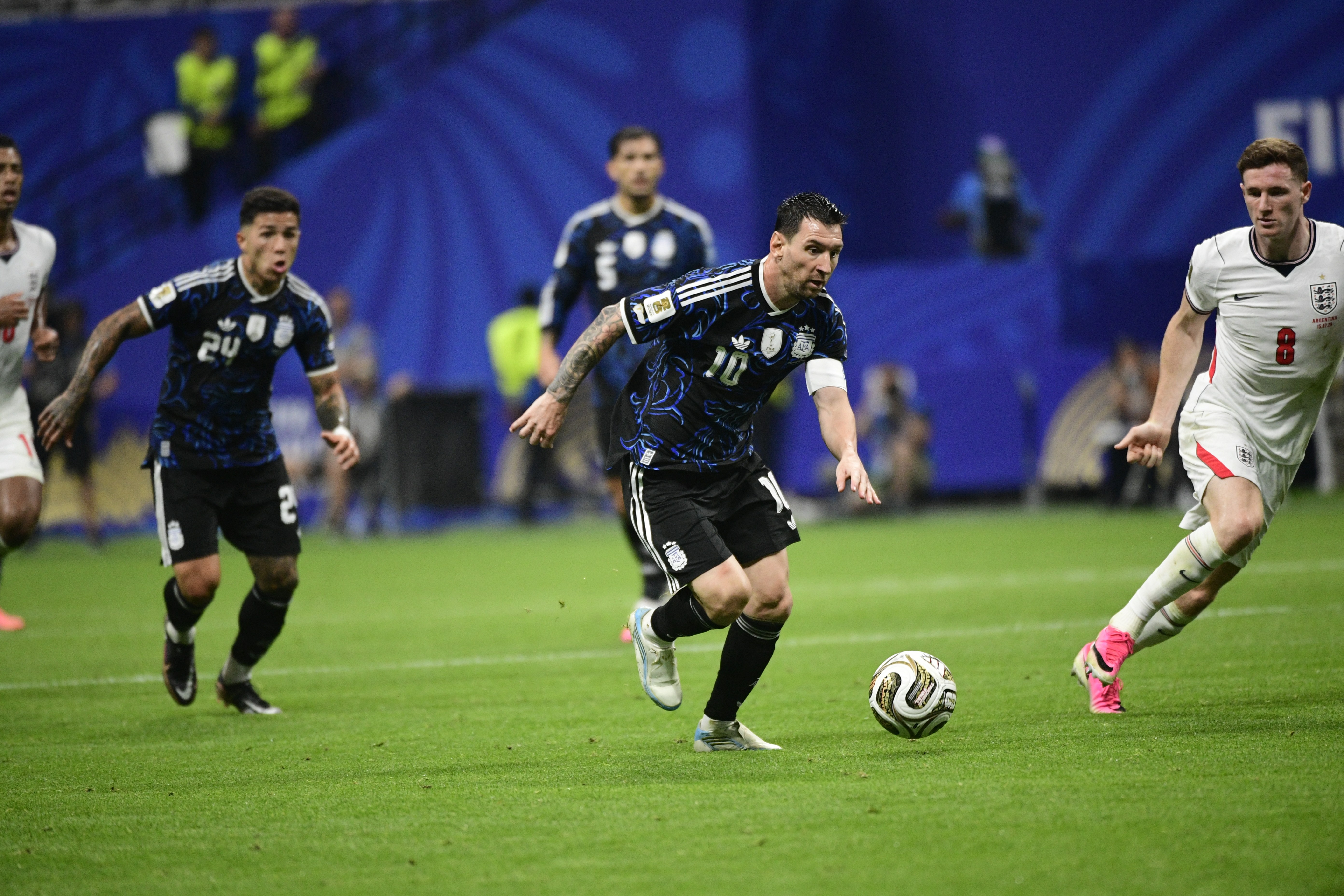 Lionel Messi, camisa 10 azul e preta, dribla a bola no campo verde, com outros jogadores ao fundo em um estádio de futebol