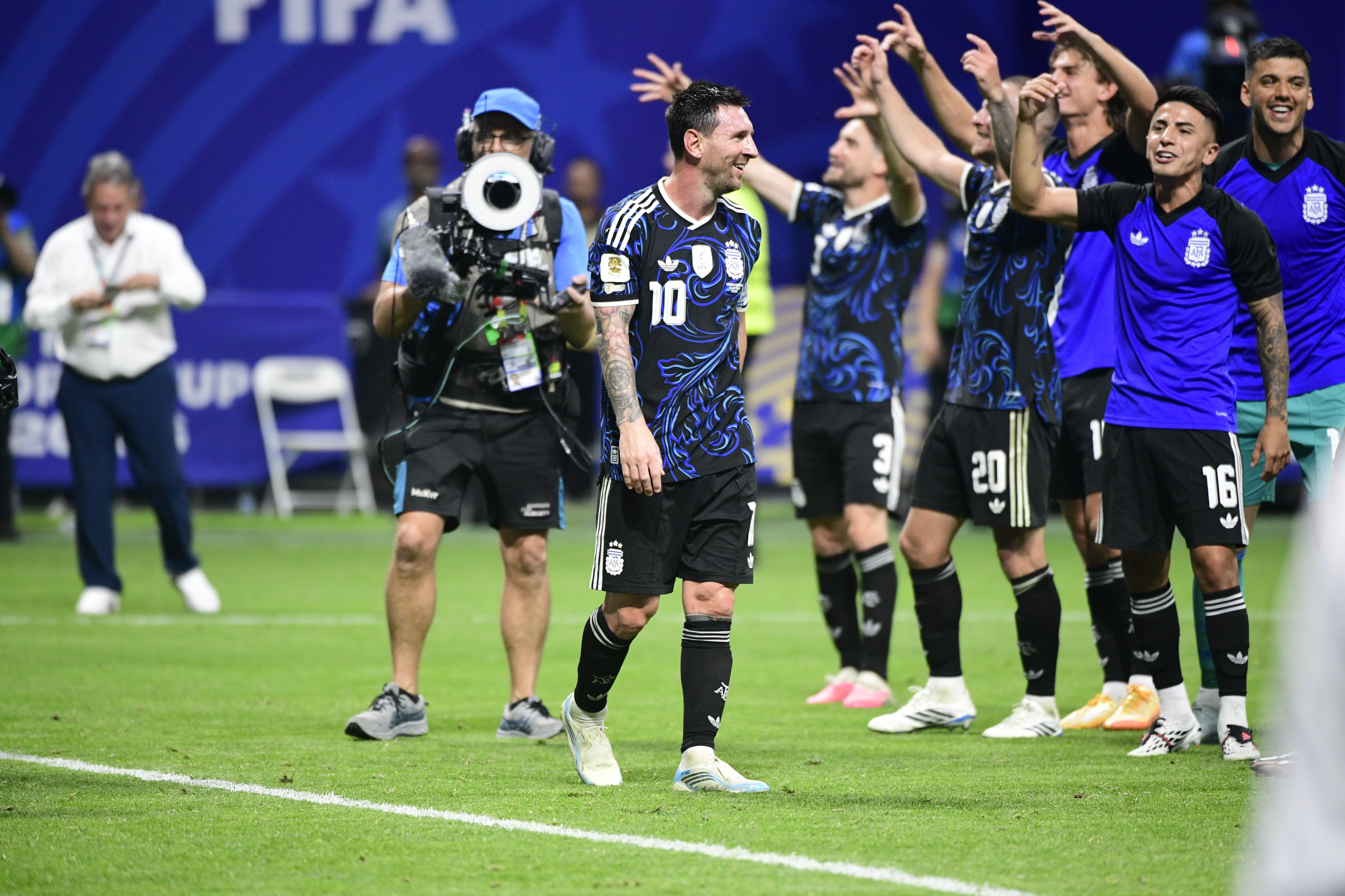 Lionel Messi, sorrindo, veste camisa preta e azul com o número 10, ao lado de outros jogadores argentinos comemorando em campo. Um cinegrafista com equipamento profissional e um homem de camisa branca e calça azul estão ao fundo, em um estádio de futebol com gramado verde e fundo azul