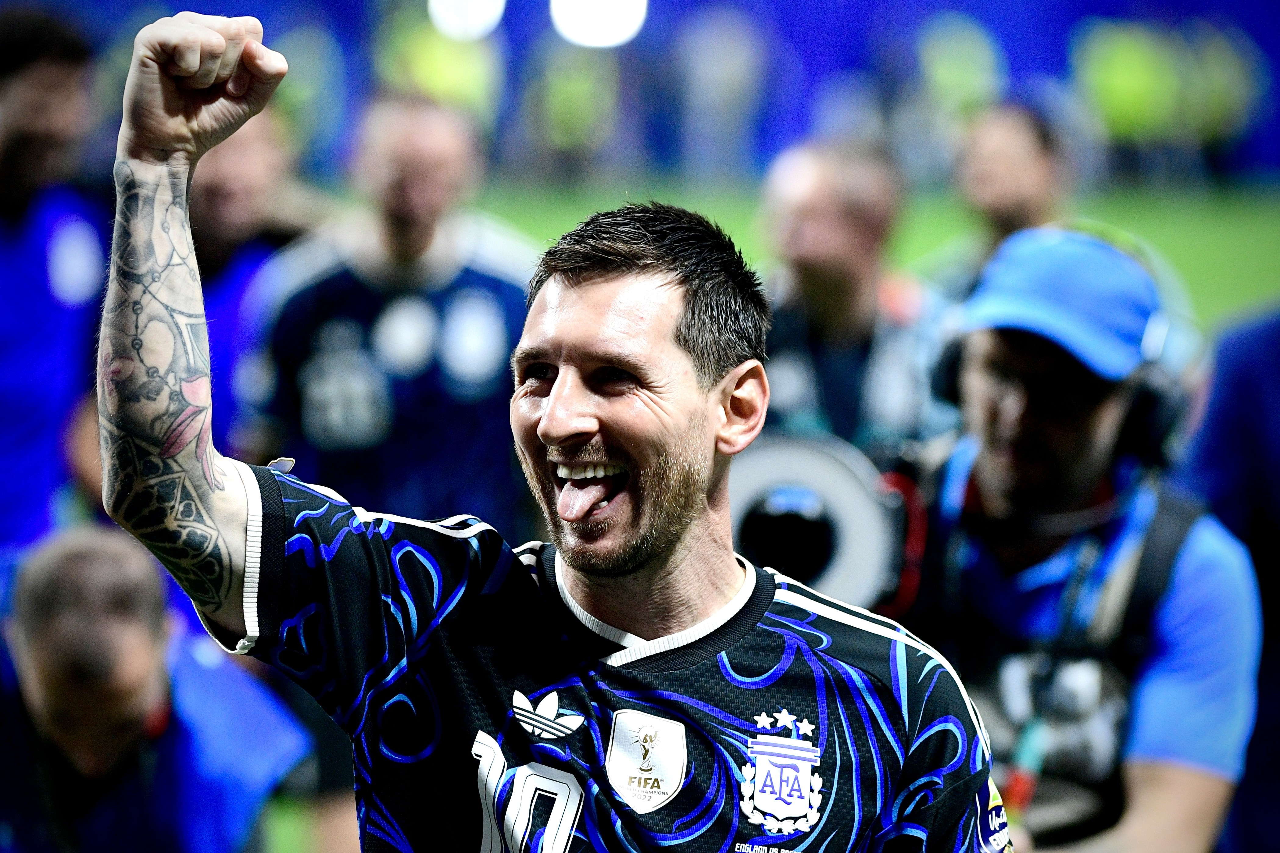 Lionel Messi, sorrindo e com a língua para fora, celebra com o punho erguido, vestindo uma camisa preta e azul com o número 10 e o escudo da FIFA