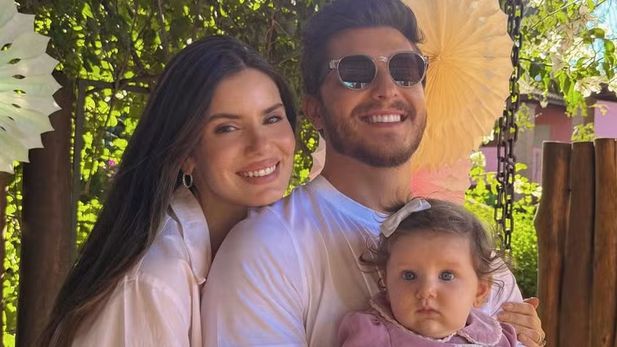 Um casal sorridente e sua bebê em um ambiente externo com folhagens e decorações de papel. A mulher, de cabelos escuros, apoia a cabeça no ombro do homem, que usa óculos de sol. A bebê, com laço branco na cabeça e roupa rosa, está no colo do pai, olhando para frente com expressão séria