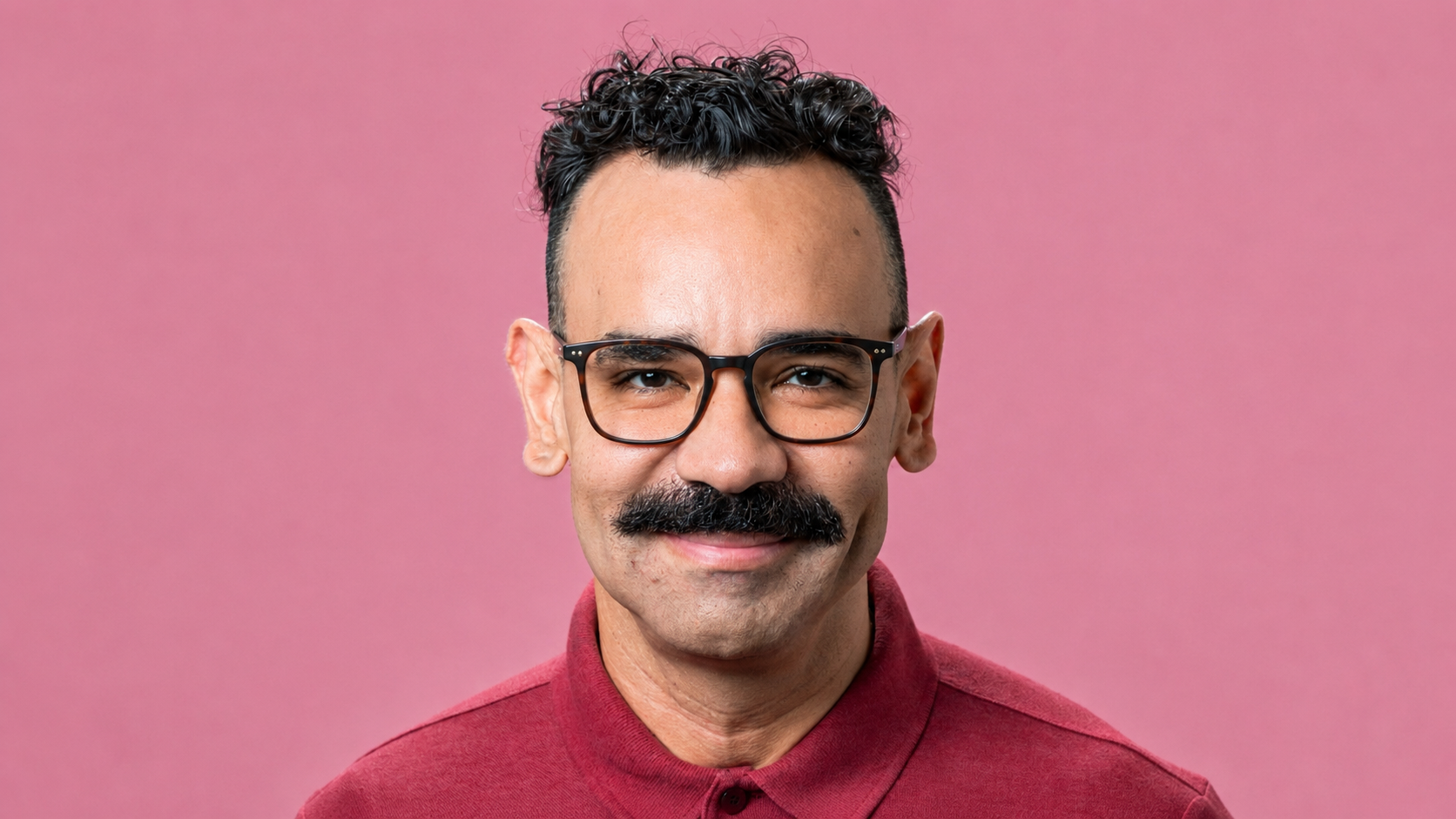 Homem de pele morena, cabelo crespo e curto, bigode volumoso, óculos de armação preta, sorrindo levemente e vestindo camisa polo vinho, em fundo rosa
