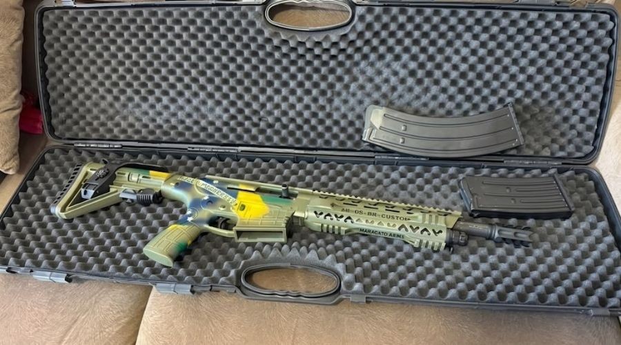 Um rifle de assalto camuflado em tons de verde, amarelo e azul, com dois carregadores, dentro de um estojo preto acolchoado com espuma, sobre um sofá marrom