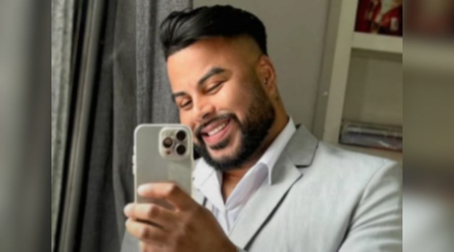 Homem sorridente, de barba e cabelo escuro bem penteado, vestindo terno cinza claro e camisa branca, tira uma selfie com um celular prateado. Ele está em um ambiente interno com cortinas escuras à esquerda e uma estante com objetos ao fundo