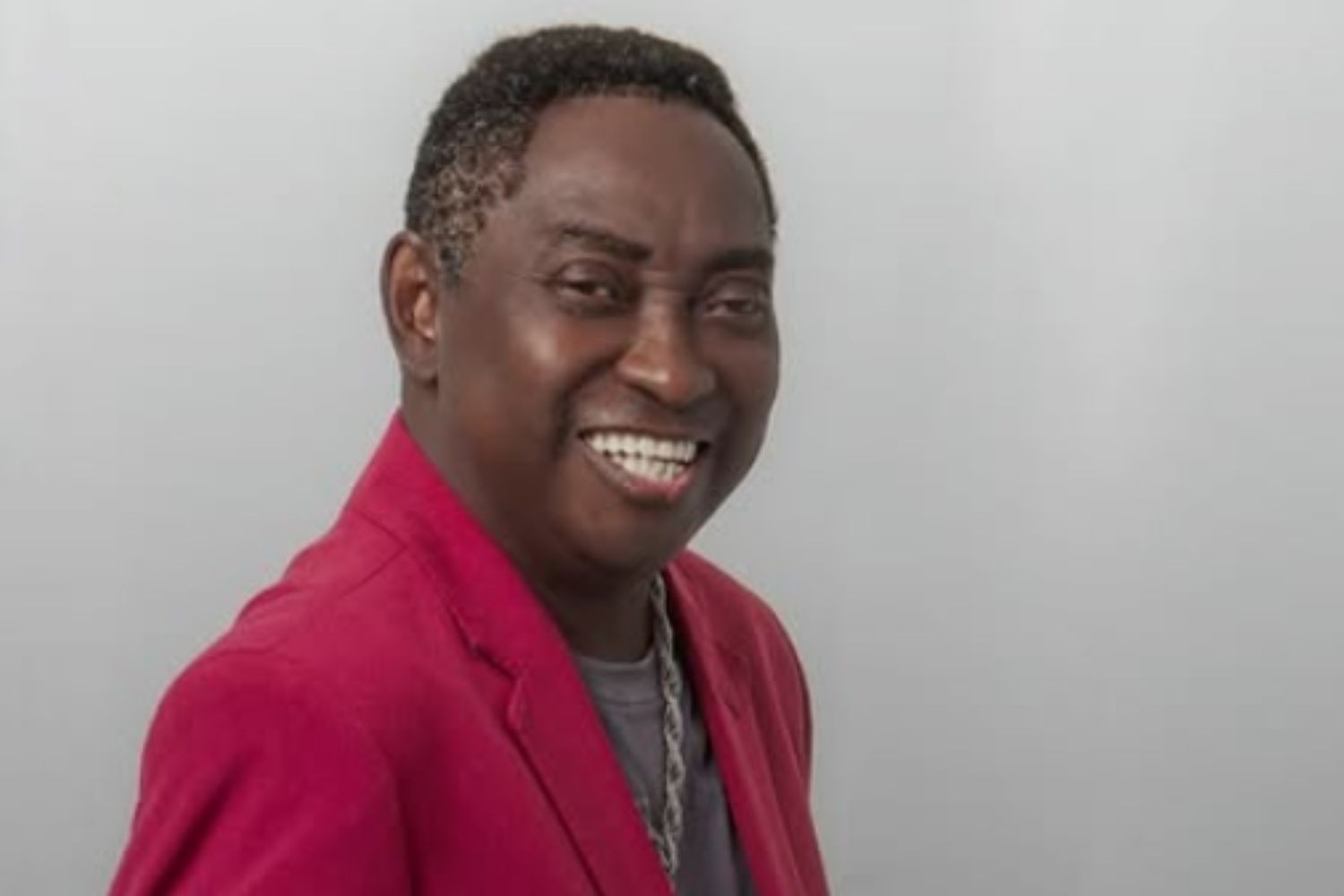 Homem negro de meia-idade, cabelo curto e crespo, sorrindo amplamente com dentes brancos, vestindo um blazer vermelho sobre uma camiseta cinza e um colar prateado, contra um fundo cinza claro