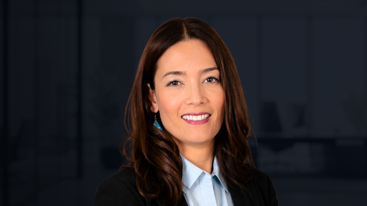Mulher de cabelos castanhos longos, sorrindo, vestindo blazer escuro e camisa social clara, com brincos azuis, em fundo de escritório desfocado