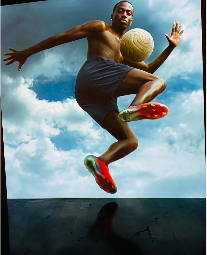 Homem negro sem camisa, usando shorts escuros e chuteiras vermelhas, salta no ar com uma bola de futebol amarela perto do peito, contra um céu azul com nuvens brancas. Sua sombra é projetada em uma superfície escura abaixo.