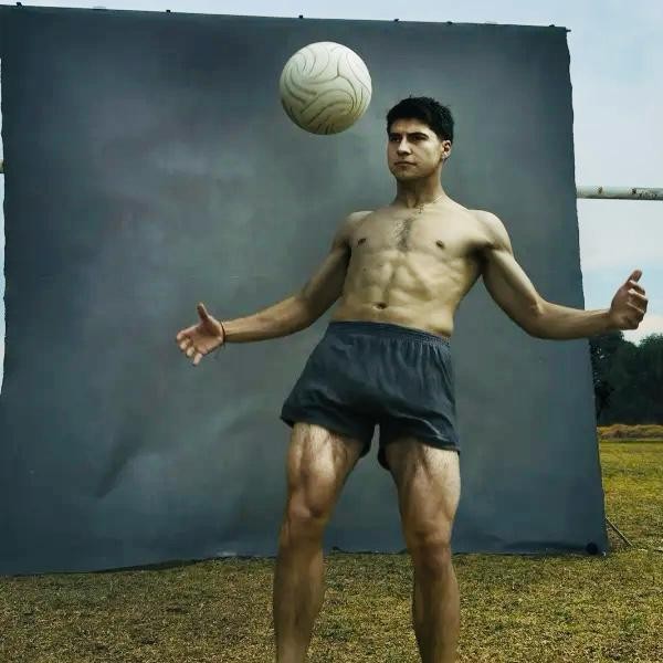 Homem sem camisa, com shorts escuros, equilibrando uma bola de futebol na cabeça, braços abertos, em frente a um fundo cinza e gramado