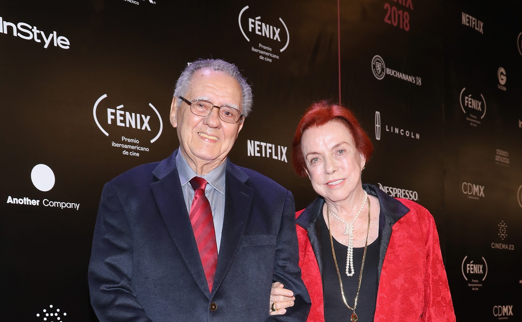 Um homem idoso de óculos e terno azul-marinho com gravata vermelha sorri ao lado de uma mulher idosa de cabelo ruivo curto, blusa preta, casaco vermelho e colares de pérolas, ambos em um evento com fundo preto e logotipos de patrocinadores como Fénix e Netflix