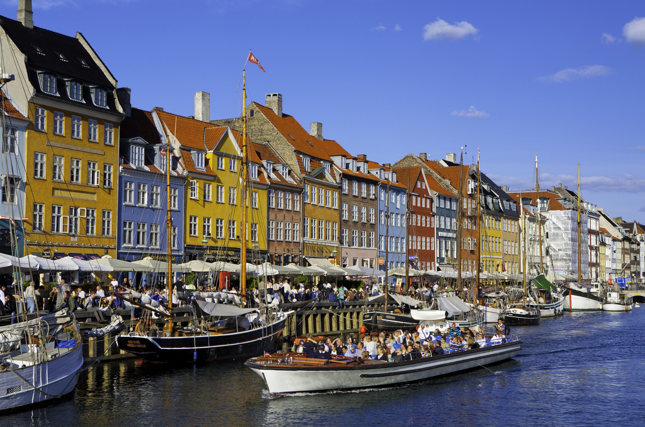 Barcos de passeio e veleiros atracados em um canal, com edifícios coloridos de vários andares e telhados inclinados ao fundo. Pessoas sentadas em mesas com guarda-sóis brancos na margem, e um barco de turismo cheio de passageiros navegando no canal. O céu é azul com poucas nuvens brancas