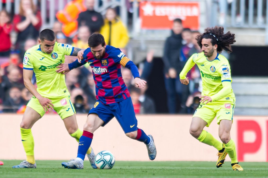Três jogadores de futebol em campo. Um jogador do Barcelona, com camisa azul e grená, dribla a bola entre dois adversários do Getafe, de uniforme verde-limão. O jogador do Barcelona está no centro, com a bola aos pés, enquanto os outros dois o marcam de perto. O fundo mostra a arquibancada com torcedores desfocados