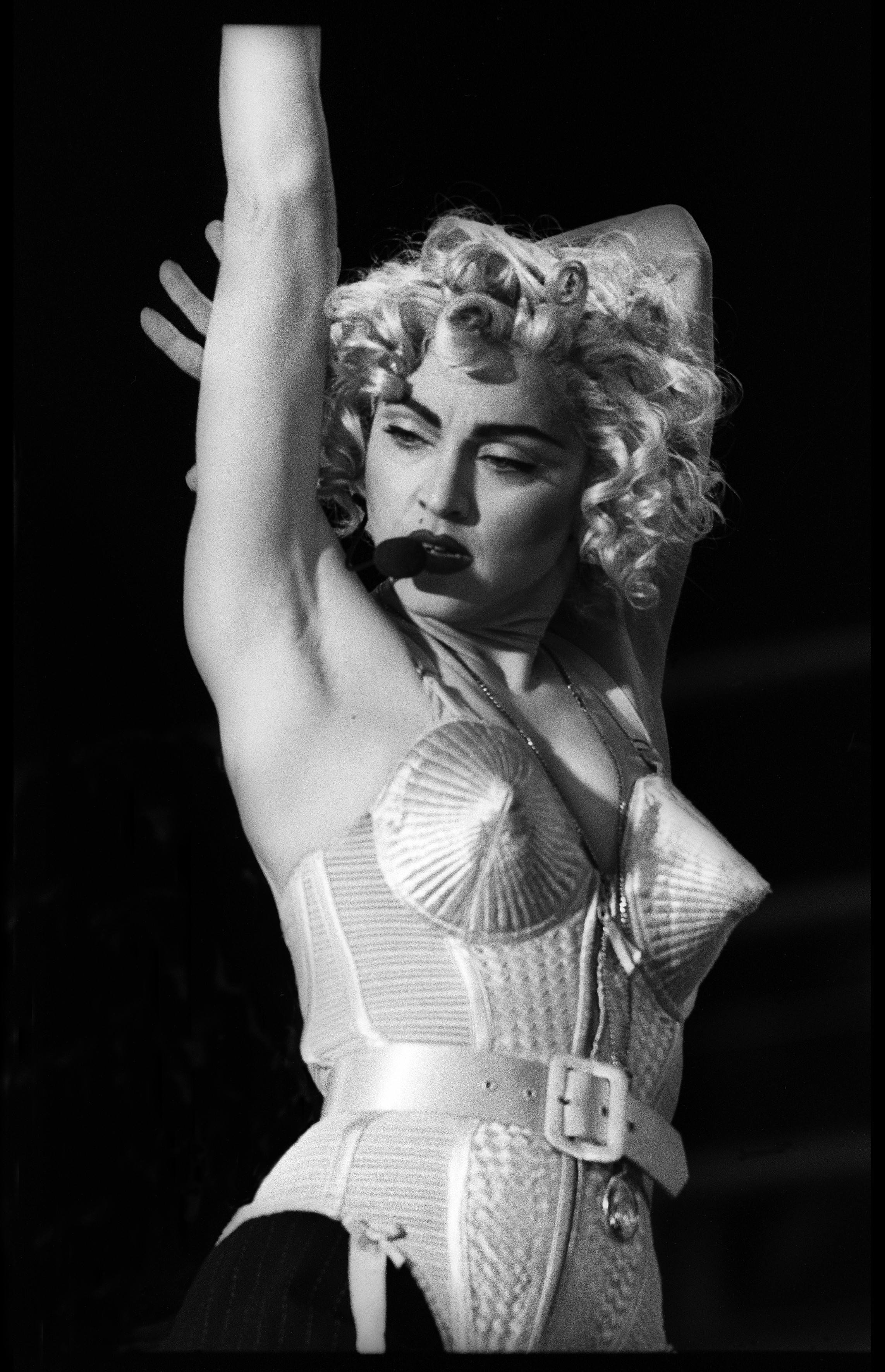 Madonna com cabelos loiros cacheados, maquiagem escura e microfone na boca, vestindo um espartilho c&ocirc;nico branco com cinto, bra&ccedil;o esquerdo levantado, em preto e branco
