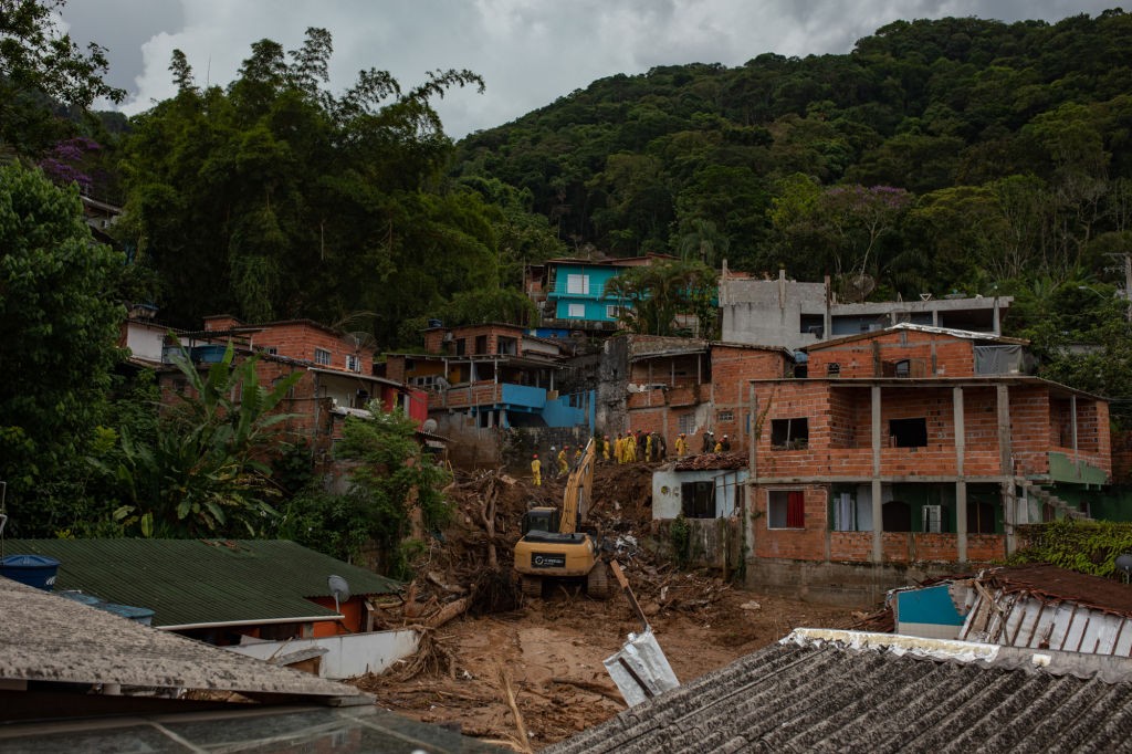 Vista aérea de uma área residencial em encosta, com casas de alvenaria e telhados variados, algumas parcialmente destruídas por um deslizamento de terra. Uma escavadeira amarela e equipes de resgate com coletes amarelos trabalham na remoção de lama e detritos. O céu está nublado e a vegetação densa cobre o topo da montanha