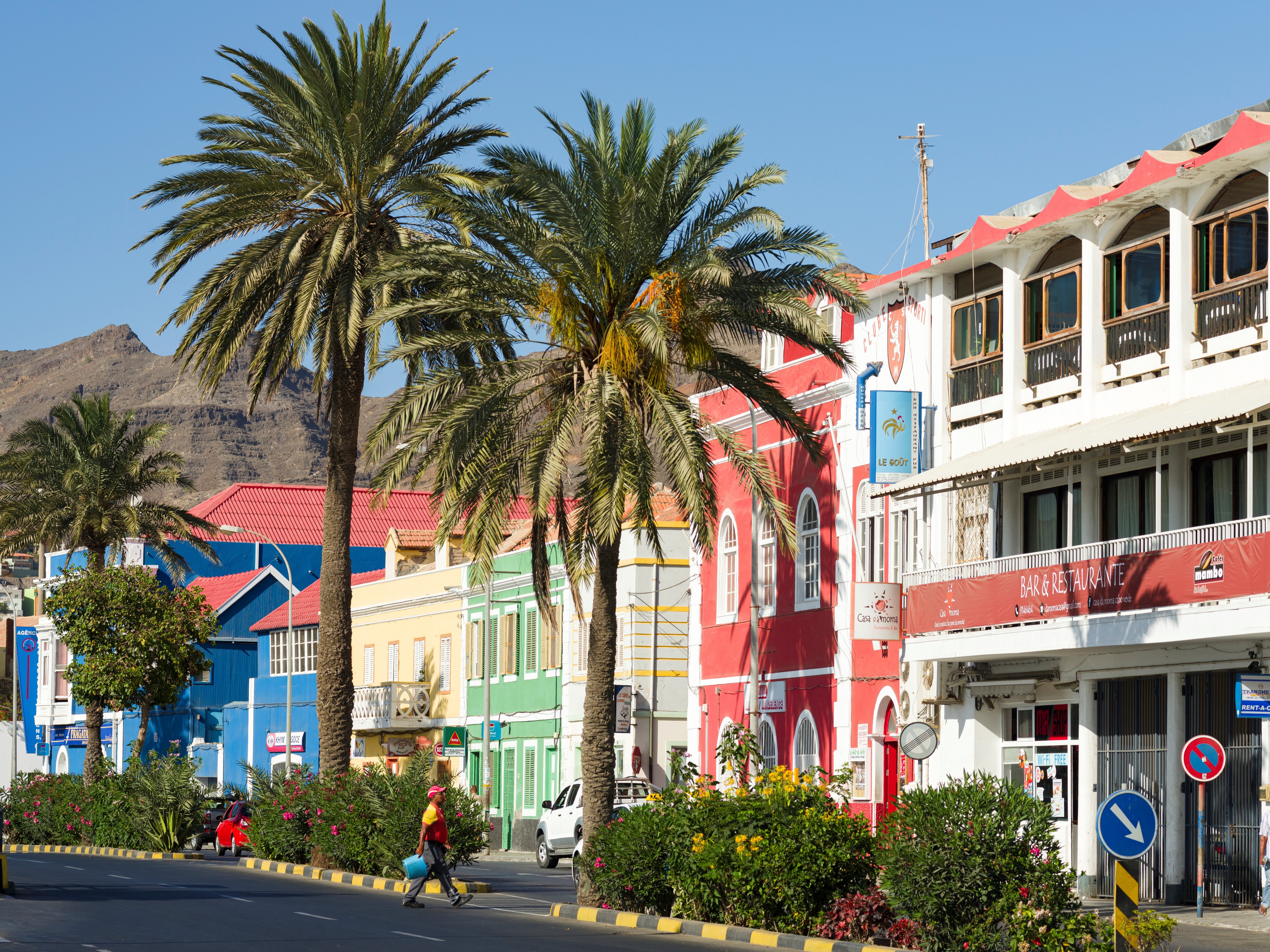 Rua colorida em Cabo Verde com palmeiras altas, edif&iacute;cios vibrantes em vermelho, azul, amarelo e verde, e montanhas ao fundo. Uma pessoa caminha na cal&ccedil;ada, e carros est&atilde;o estacionados.