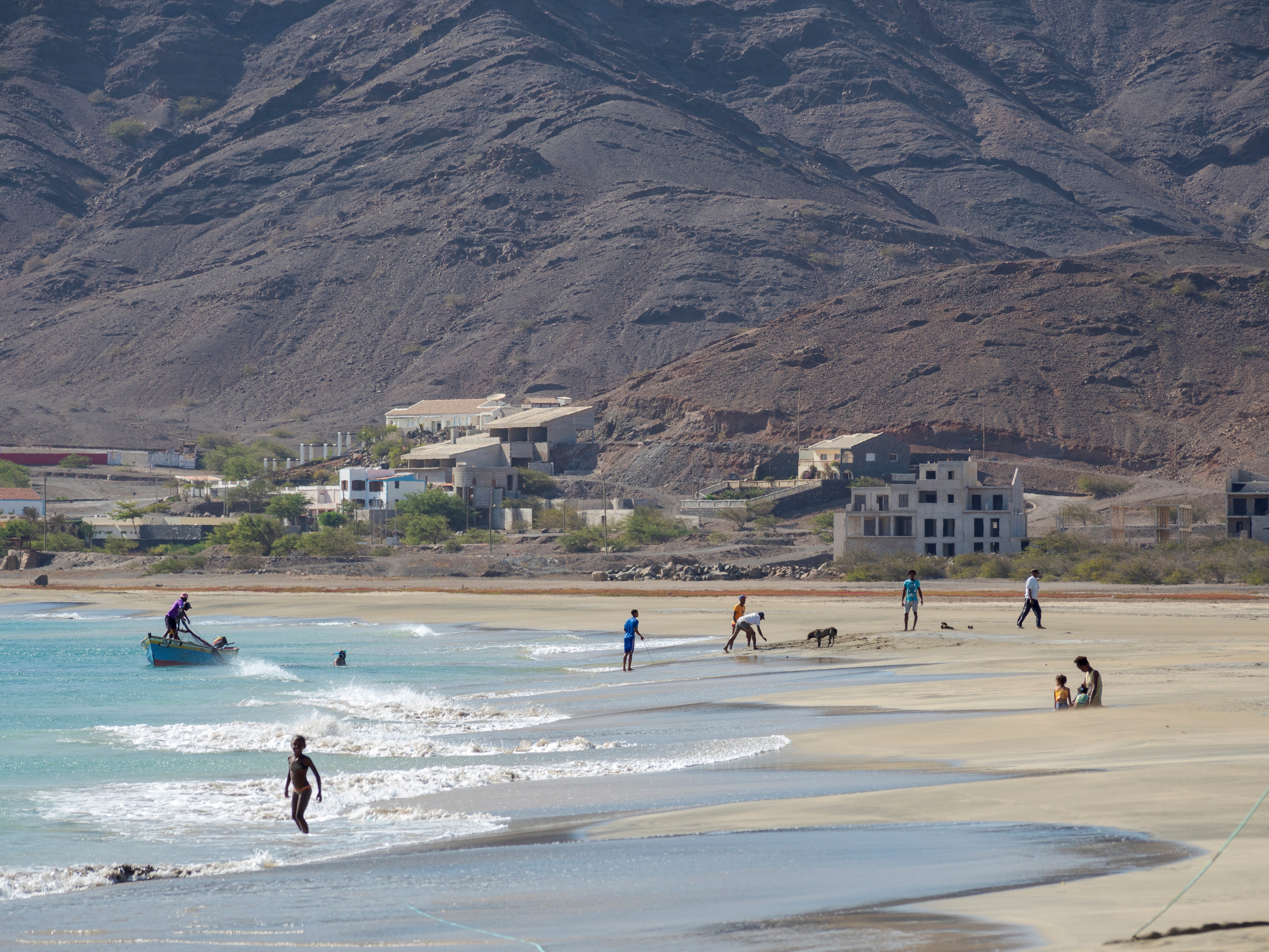 Pessoas na praia de areia clara com mar azul-turquesa, ondas pequenas e montanhas rochosas ao fundo. Uma crian&ccedil;a corre na &aacute;gua, um homem em um barco, e outros adultos e crian&ccedil;as caminham ou brincam na areia. Casas e pr&eacute;dios de cor clara est&atilde;o na encosta da montanha