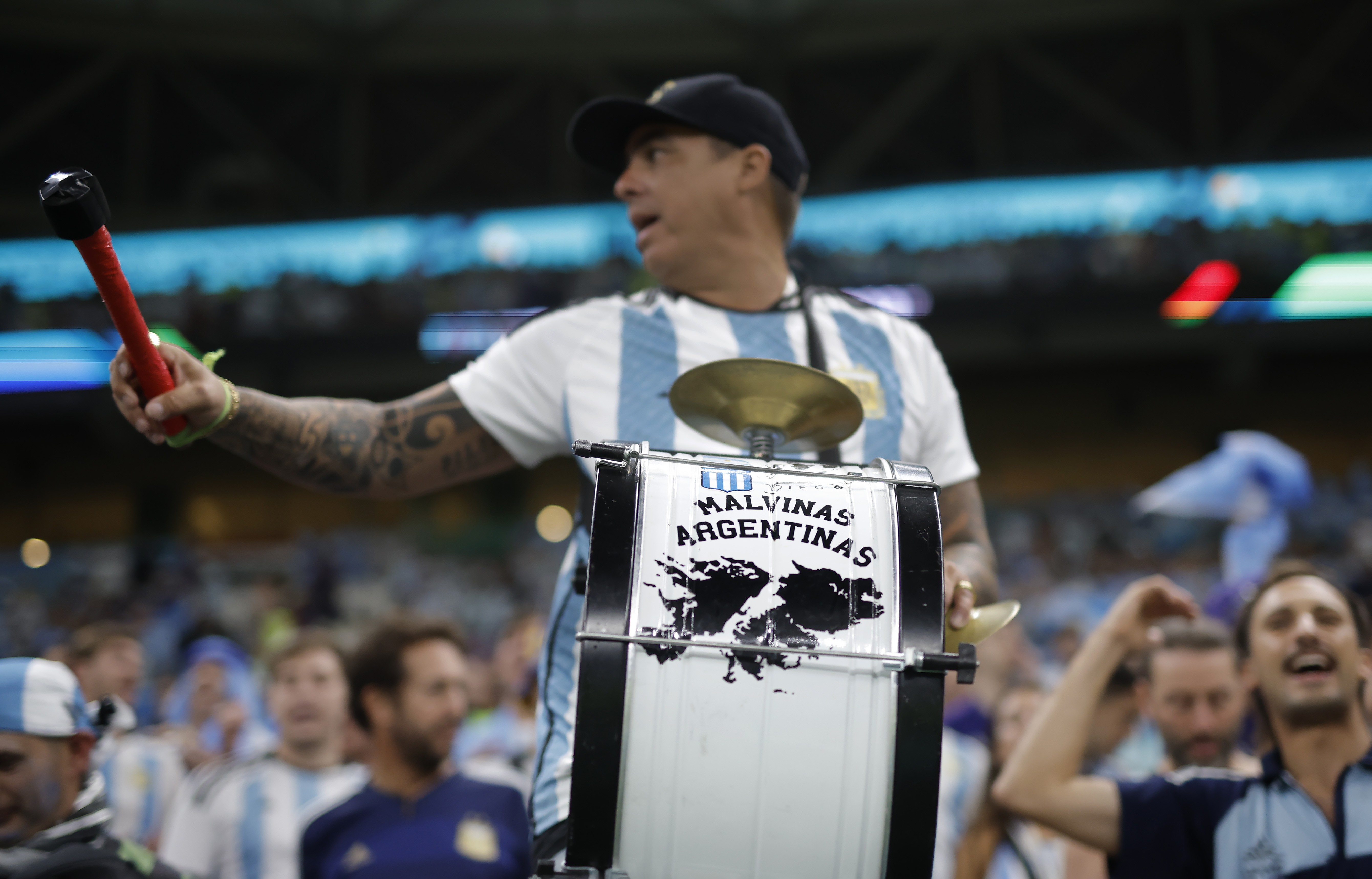 Torcedor argentino com boné preto e camisa listrada azul e branca, tocando um tambor com a frase Malvinas Argentinas e segurando um bastão vermelho, em um estádio lotado