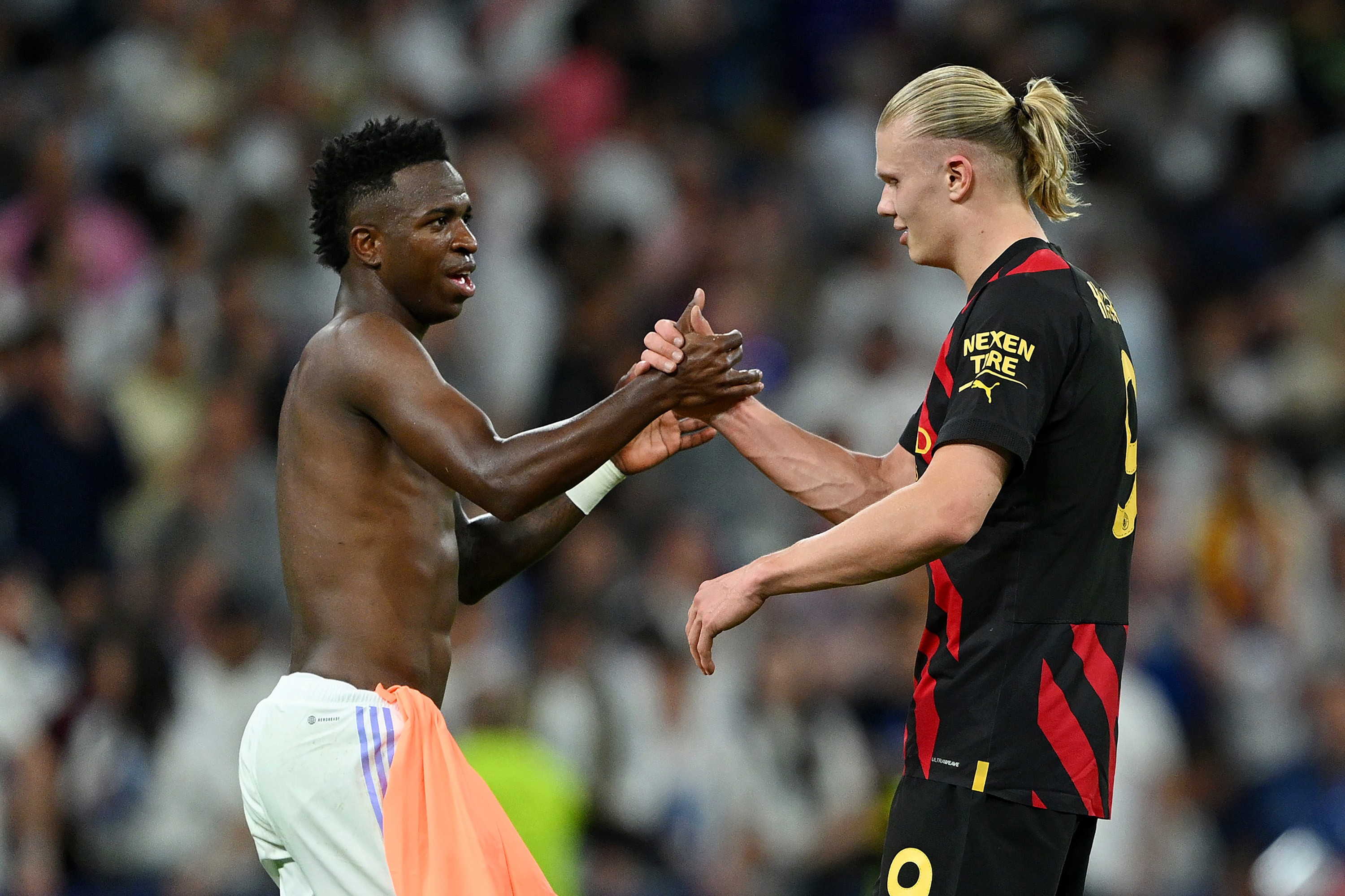 Dois jogadores de futebol, um homem negro sem camisa e outro branco de camisa preta e vermelha, apertam as mãos em campo, com torcedores borrados ao fundo