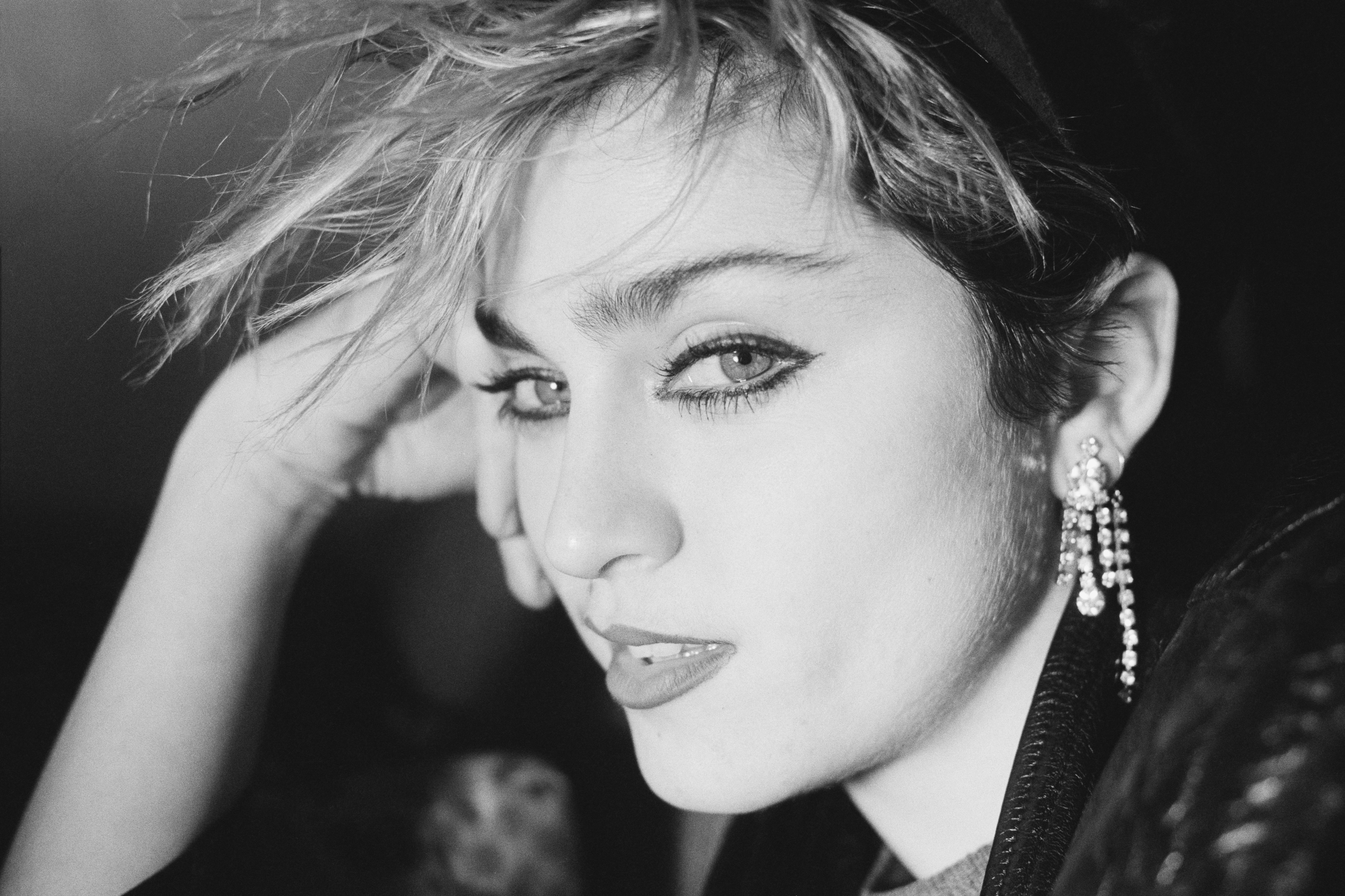 Retrato em preto e branco de Madonna jovem, com cabelo loiro esvoa&ccedil;ante, maquiagem marcante nos olhos e um brinco longo de strass na orelha direita. Ela apoia a m&atilde;o esquerda na cabe&ccedil;a e olha diretamente para a c&acirc;mera com uma express&atilde;o s&eacute;ria