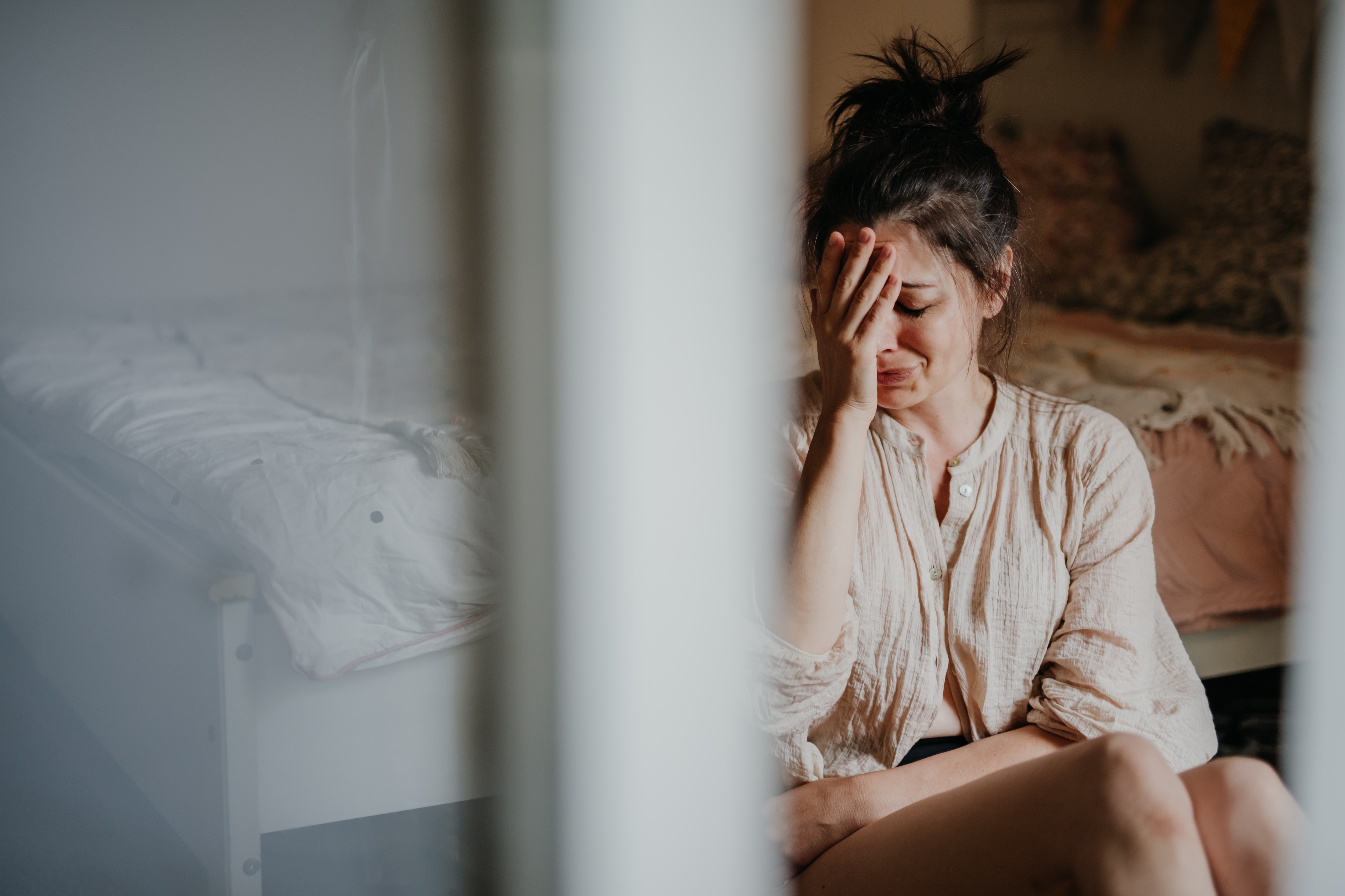 Mulher de cabelo preso, camisa bege, sentada no chão, chorando com a mão no rosto, em um quarto com cama ao fundo