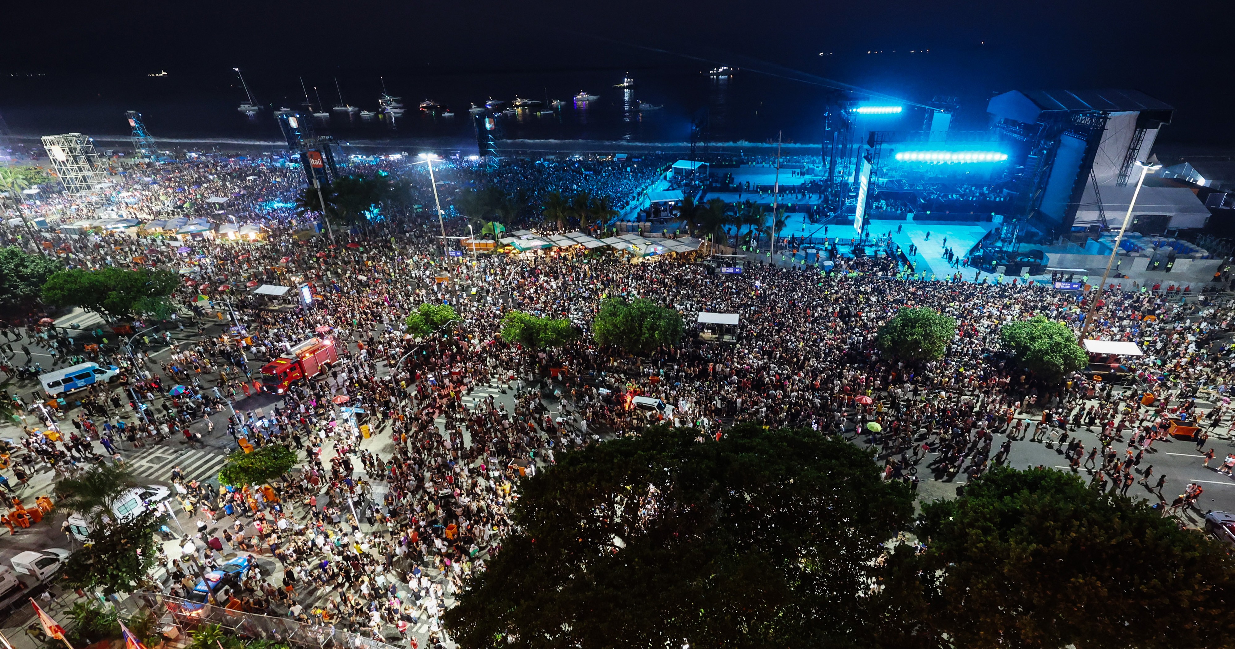 Multid&atilde;o de pessoas em um evento noturno ao ar livre, com um palco iluminado em azul &agrave; direita e barcos no mar ao fundo. H&aacute; &aacute;rvores escuras e ve&iacute;culos de emerg&ecirc;ncia, como um caminh&atilde;o de bombeiros vermelho, espalhados pela &aacute;rea. As ruas est&atilde;o cheias de pedestres, e a ilumina&ccedil;&atilde;o p&uacute;blica &eacute; intensa