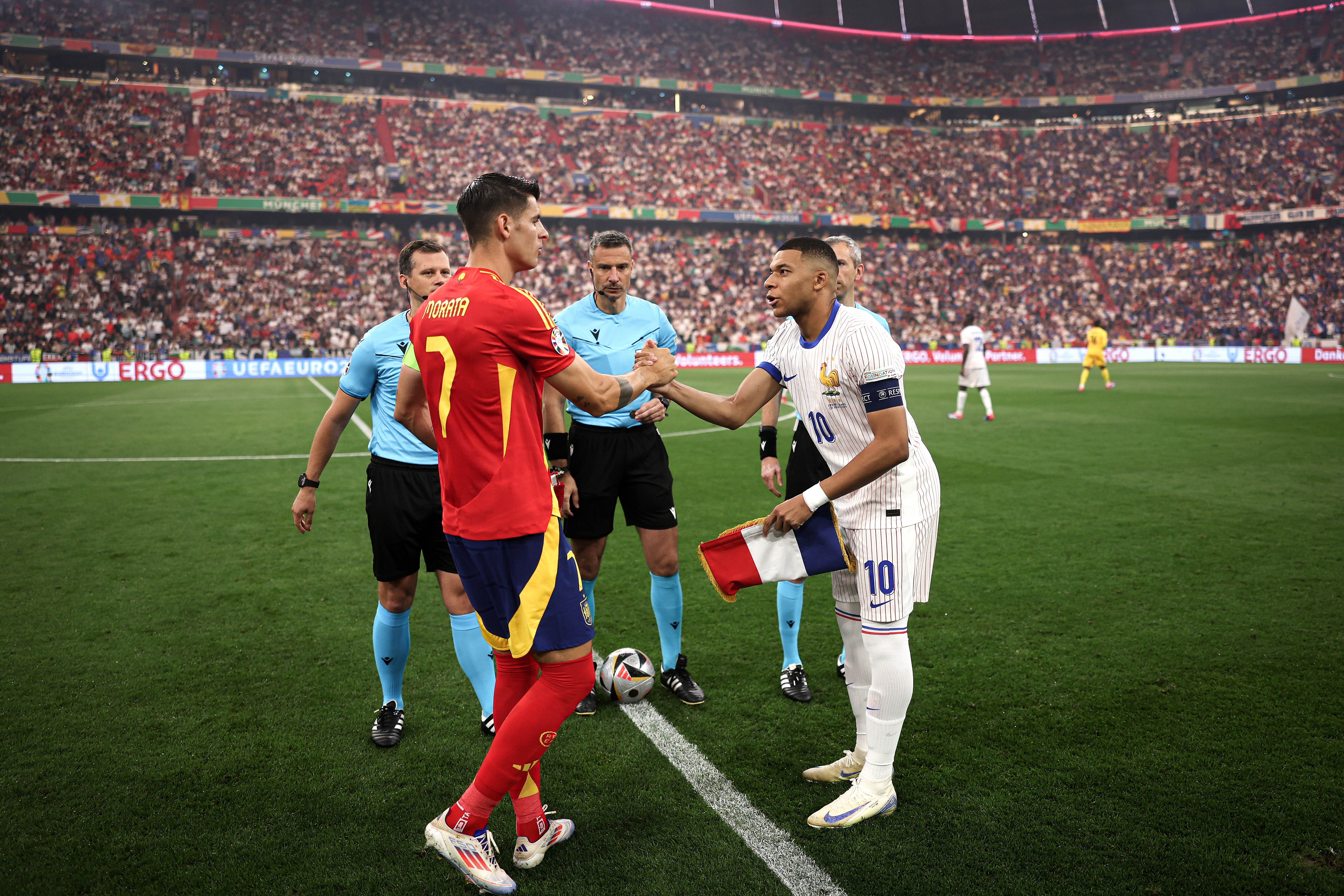 Dois jogadores de futebol, um com camisa vermelha e outro com camisa branca listrada, apertam as mãos no centro de um campo de futebol, com árbitros e um estádio lotado ao fundo