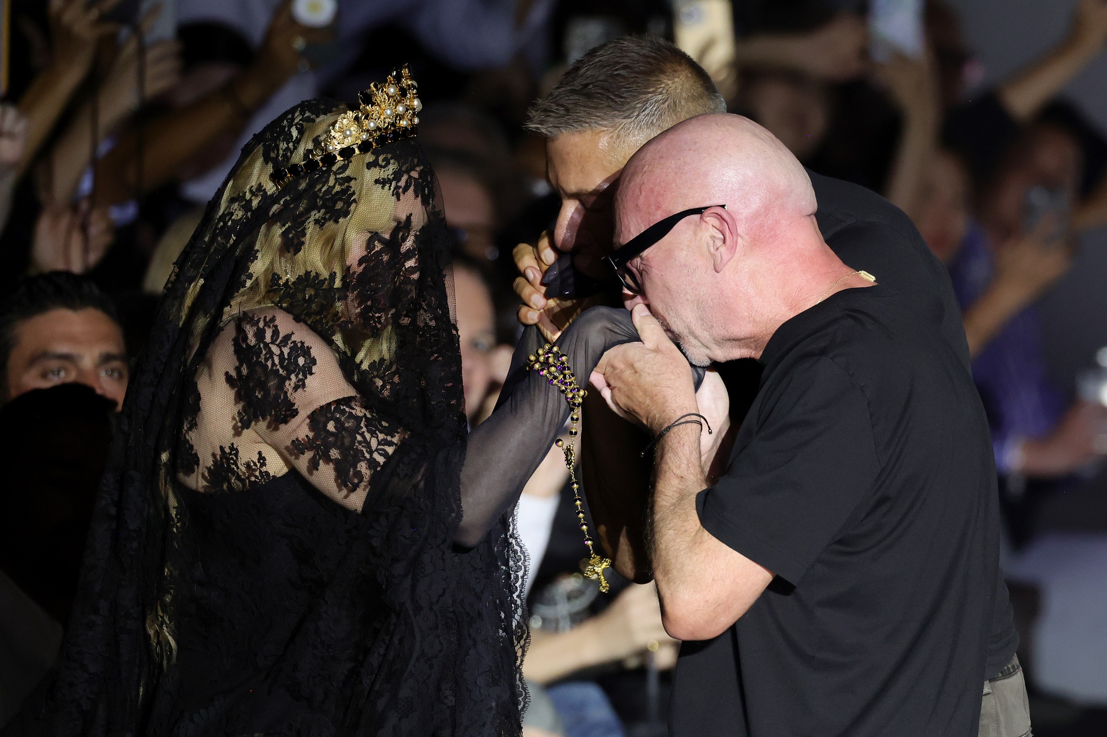 Uma mulher loira com v&eacute;u preto de renda e coroa dourada na cabe&ccedil;a, vestindo luvas de renda, segura um ter&ccedil;o dourado enquanto um homem careca de &oacute;culos escuros e camiseta preta beija sua m&atilde;o. Outro homem de cabelo grisalho observa ao fundo.