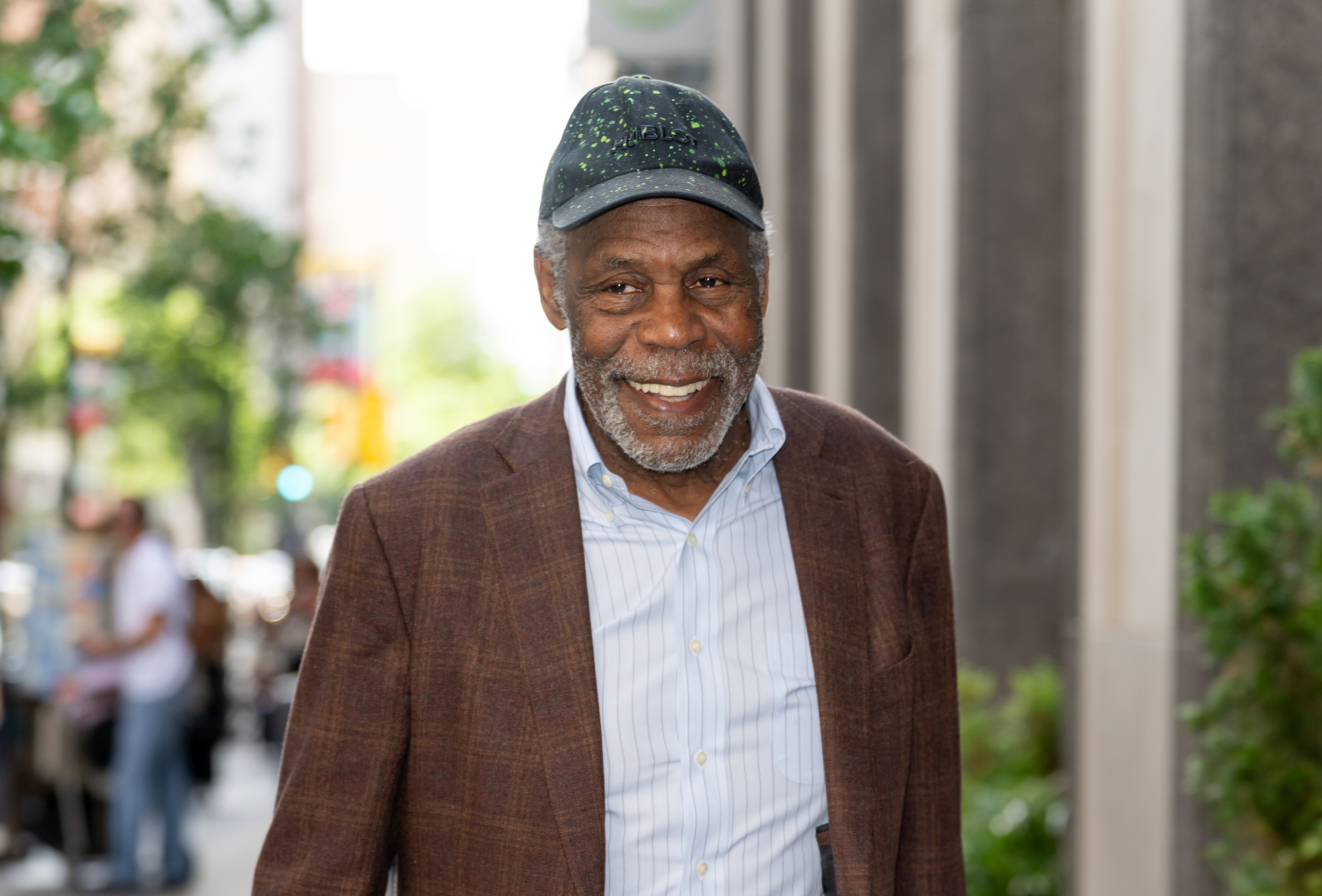 Homem negro idoso, com barba e bigode grisalhos, sorrindo e olhando para frente. Veste boné escuro com manchas claras, camisa social azul clara listrada e blazer marrom. Ao fundo, uma rua com pessoas borradas e um prédio com pilares verticais