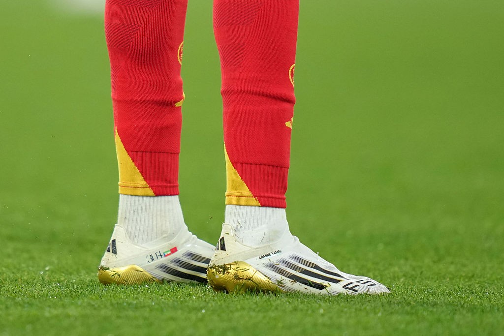 Pés de um jogador de futebol em campo, usando meias vermelhas com detalhes amarelos, meias brancas e chuteiras brancas com solado dourado e listras pretas, sobre a grama verde