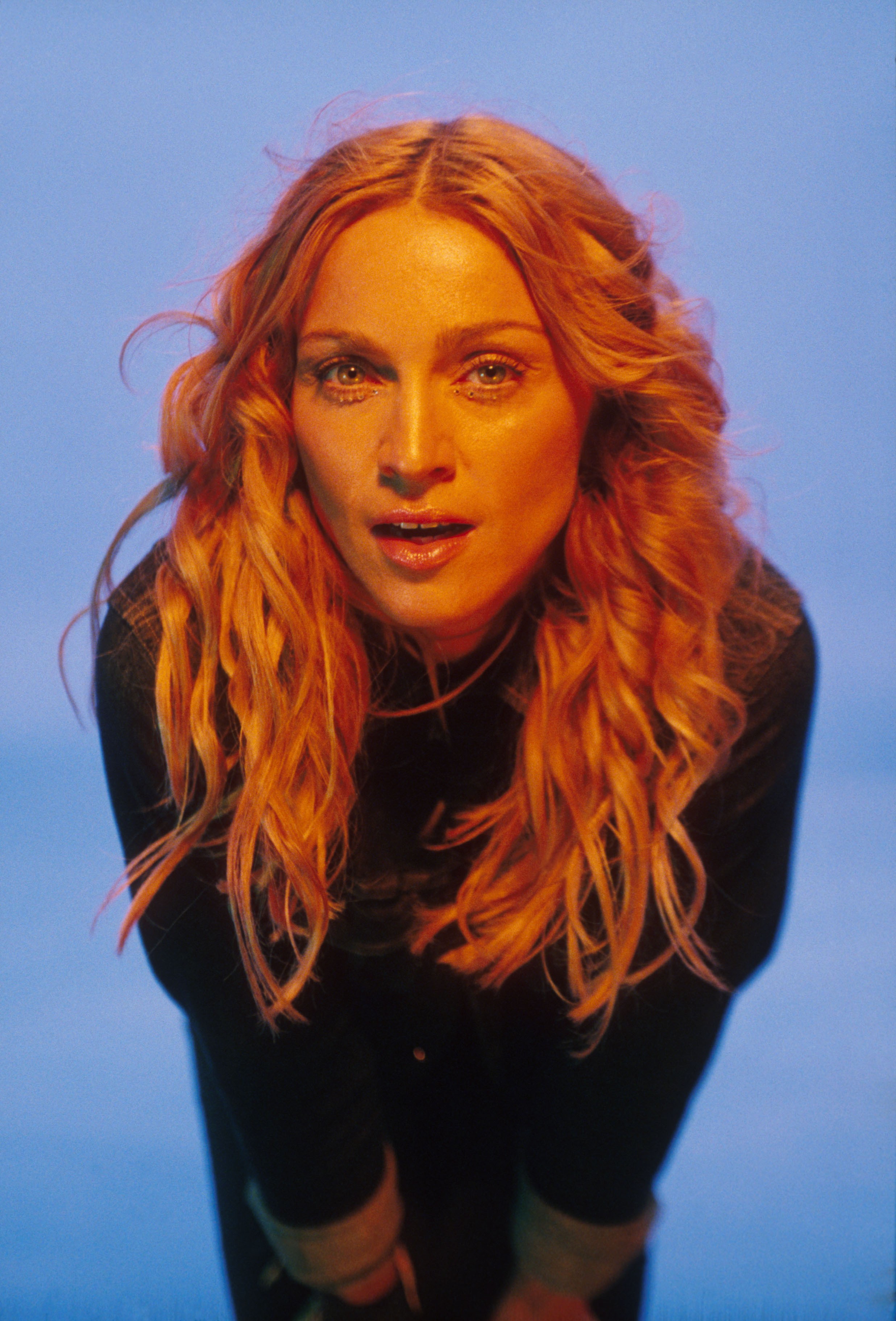 Madonna, com cabelos loiros avermelhados e ondulados, olhando diretamente para a c&acirc;mera com a boca ligeiramente aberta. Ela veste uma blusa preta de gola alta, com a pele do rosto e pesco&ccedil;o iluminada por uma luz quente, contrastando com o fundo azul