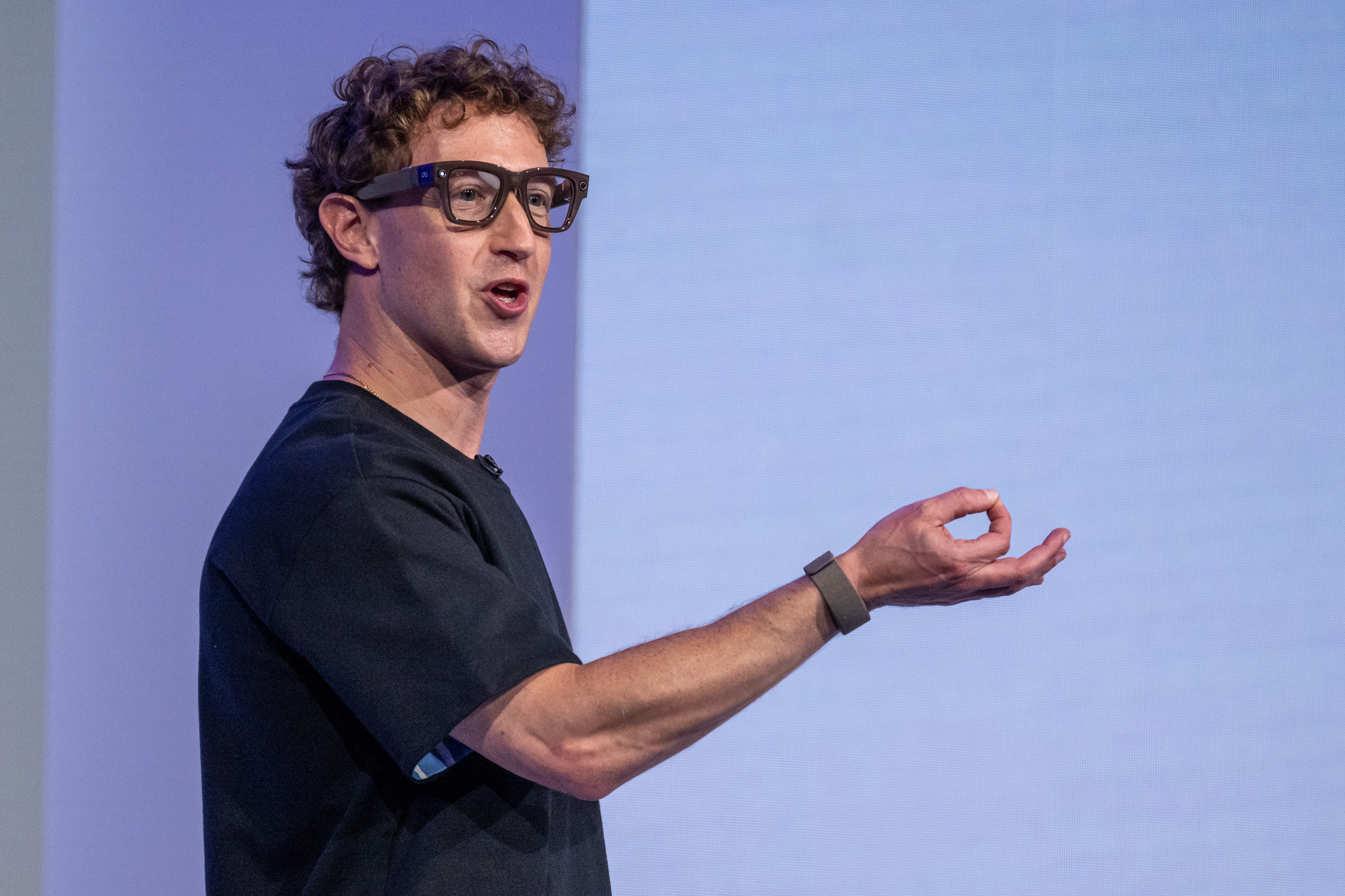 Mark Zuckerberg, com cabelo cacheado e óculos escuros, fala com a boca aberta e gesticula com a mão direita, usando uma pulseira escura, contra um fundo azul claro