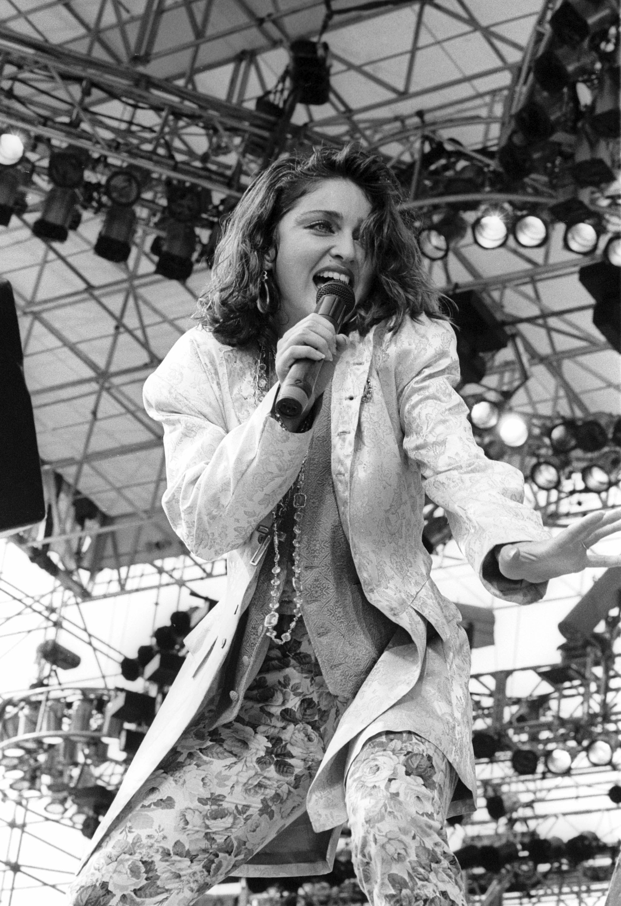 Madonna cantando em um palco, vestindo um blazer claro sobre um colete e cal&ccedil;as estampadas com flores, com colares longos e brincos grandes. Ela segura um microfone com a m&atilde;o direita, a boca aberta em express&atilde;o de canto, e a m&atilde;o esquerda estendida. O fundo mostra a estrutura met&aacute;lica do palco com luzes de show