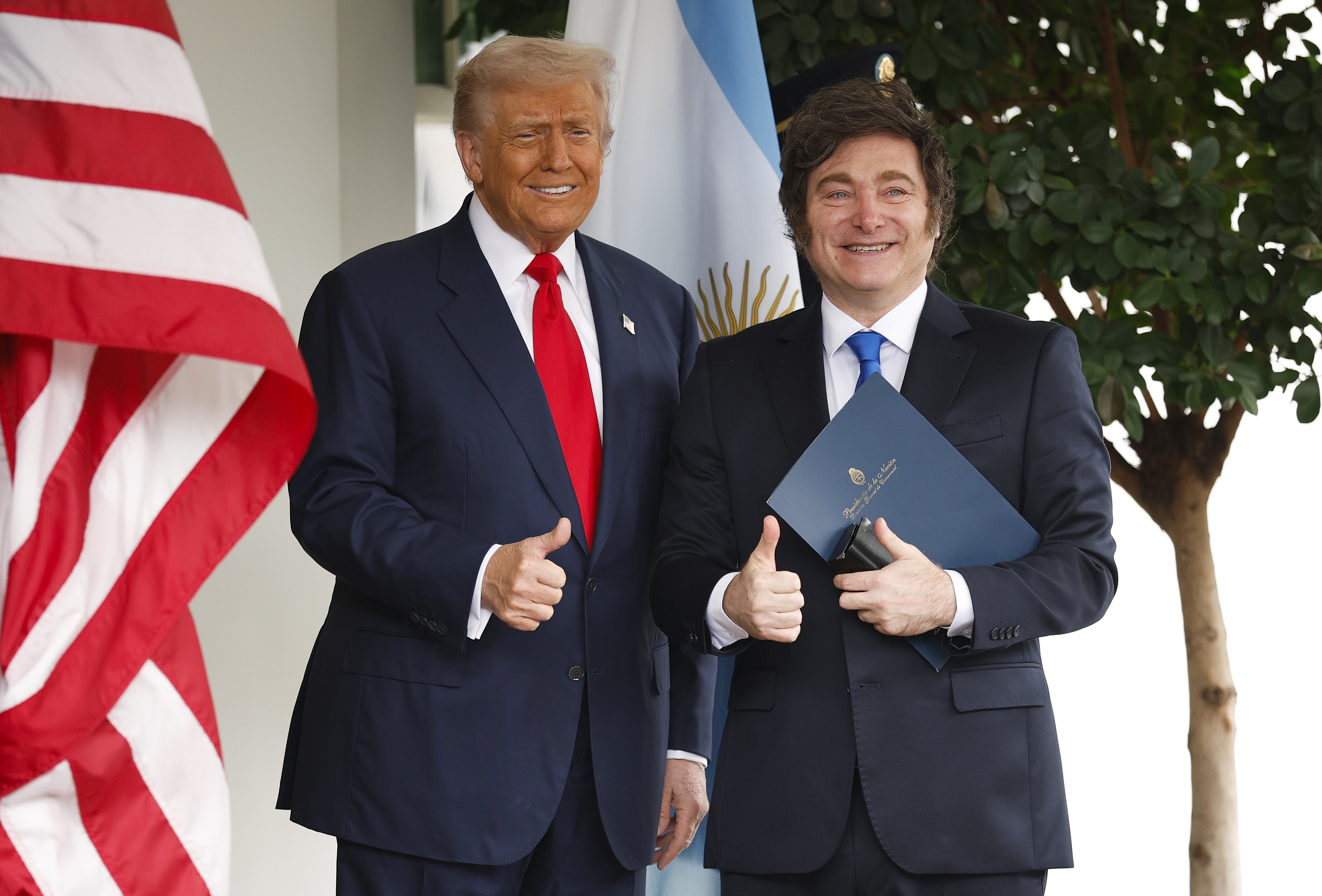 Donald Trump e Javier Milei, ambos sorridentes e de terno, posam com os polegares para cima. Trump usa terno azul-marinho e gravata vermelha, enquanto Milei veste terno preto e gravata azul, segurando uma pasta azul. Bandeiras dos EUA e Argentina ao fundo, com uma árvore à direita