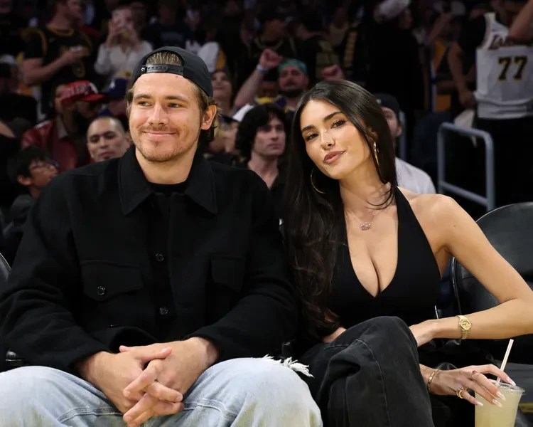 Um homem e uma mulher jovens sentados em uma arquibancada de estádio. Ele usa boné virado para trás, camisa preta e jeans claro, sorrindo levemente. Ela tem cabelos longos e escuros, usa top preto decotado, calça escura, brincos de argola e segura um copo com canudo.