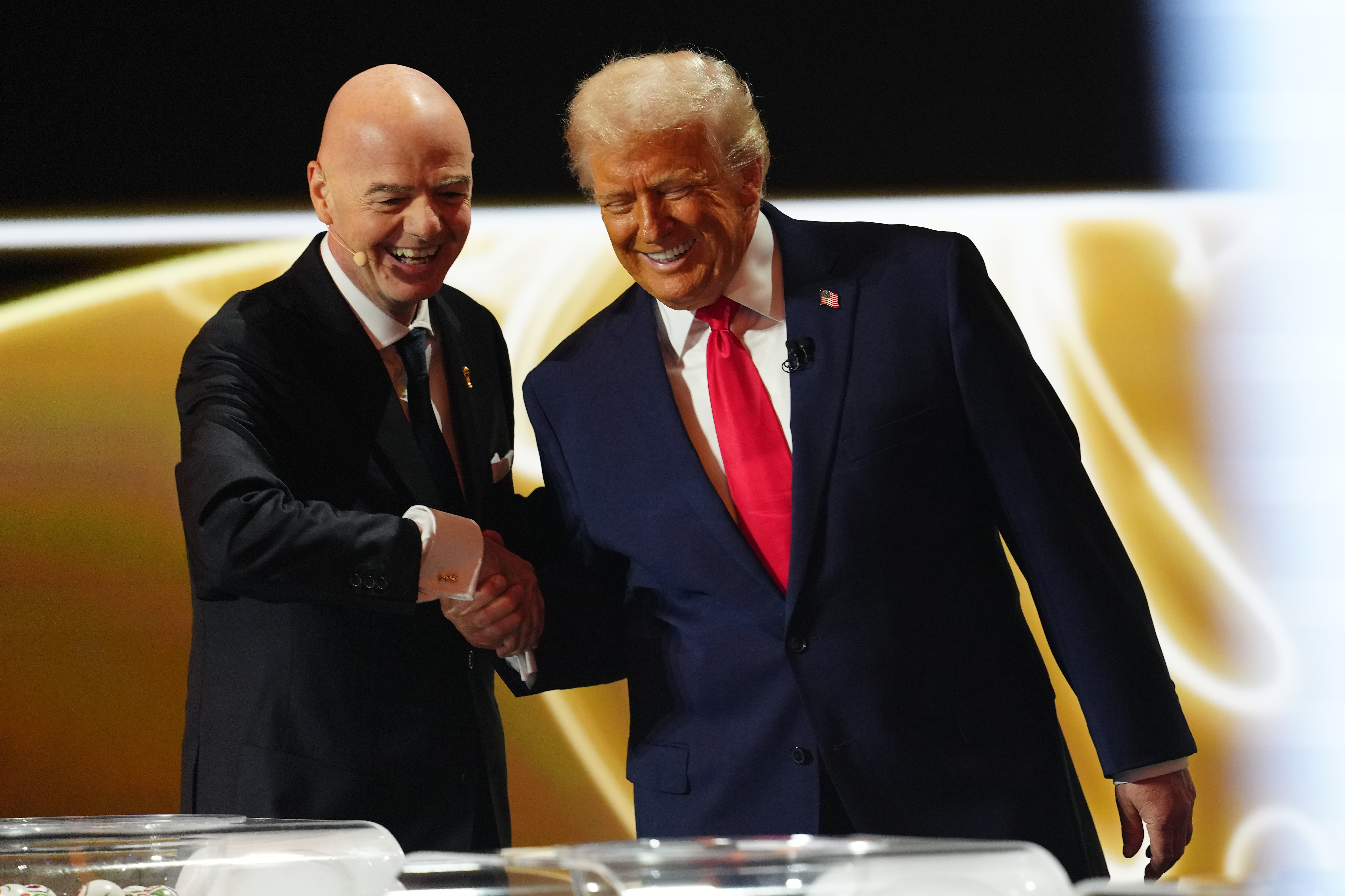 Gianni Infantino, careca, de terno preto, gravata azul e lenço branco, sorri enquanto aperta a mão de Donald Trump, de terno azul-marinho, camisa branca e gravata vermelha, que também sorri. Ambos estão em um palco com fundo dourado e branco, e há uma mesa transparente em primeiro plano
