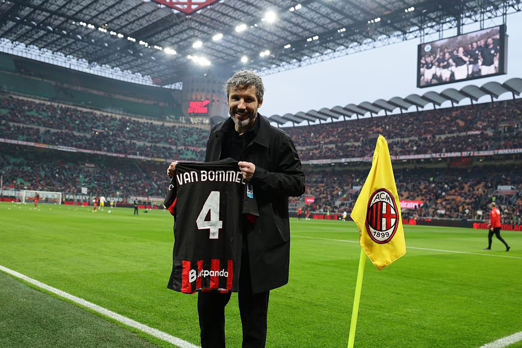 Homem de barba grisalha e casaco preto sorri, segurando uma camisa de futebol preta com VAN BOMMEL 4 e Bitpanda em um estádio lotado. Uma bandeira amarela do AC Milan está à direita, e um telão exibe imagens no fundo