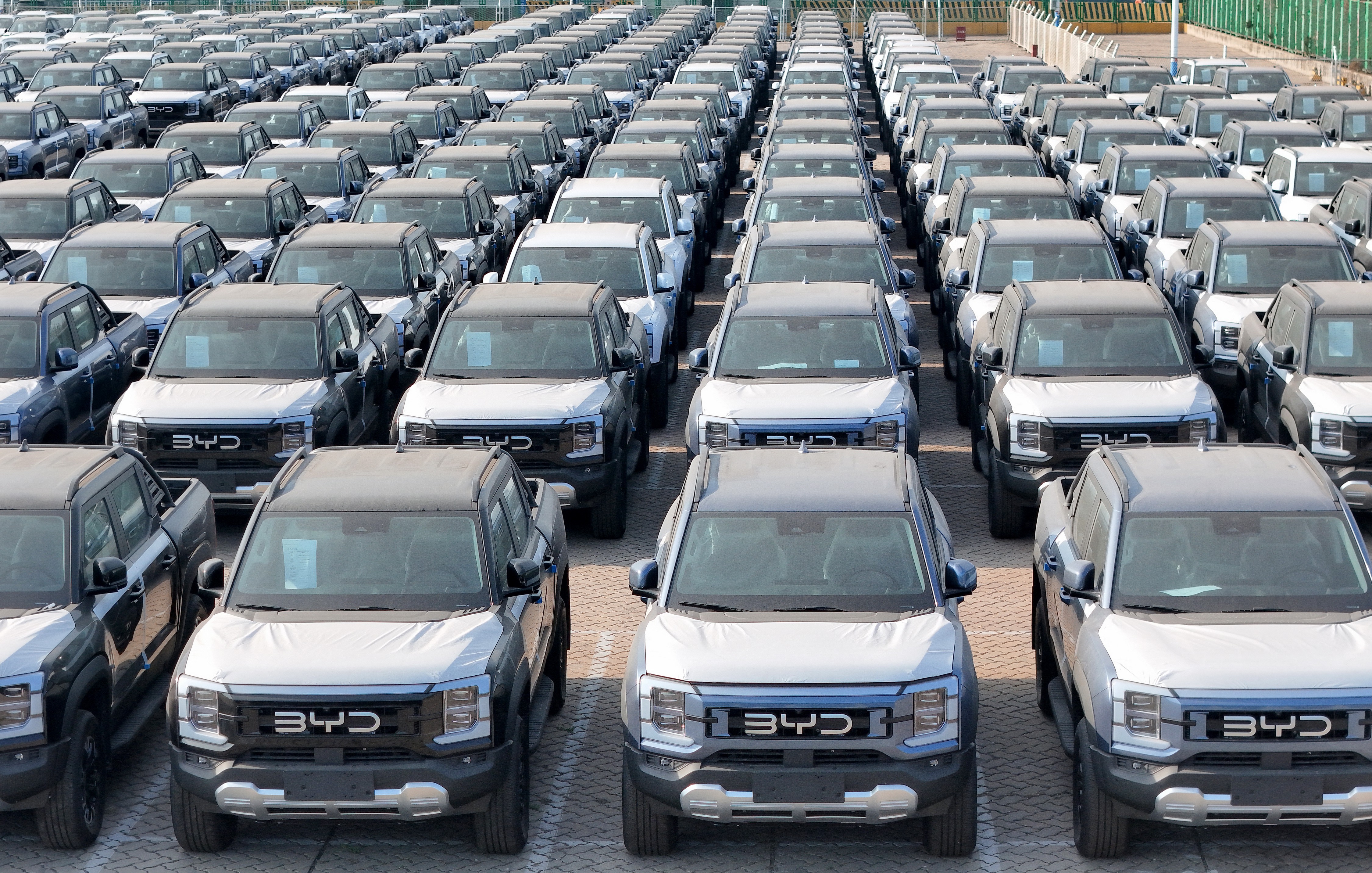 Centenas de picapes BYD Shark cinza e brancas, com capôs parcialmente cobertos, estacionadas em fileiras organizadas em um pátio de distribuição, vistas de cima