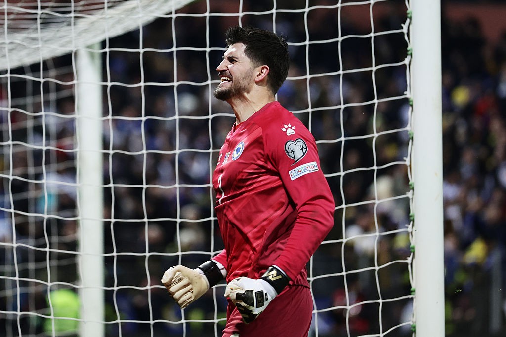 Goleiro de futebol com barba, vestindo uniforme vermelho e luvas brancas, expressa frustração com a boca aberta e os punhos cerrados, em frente à rede do gol durante uma partida noturna