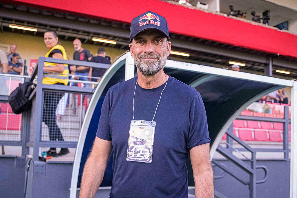 Homem de barba grisalha, boné Red Bull e camiseta azul marinho, com crachá no pescoço, sorri para a câmera em um estádio de futebol. Ao fundo, pessoas e arquibancadas vermelhas