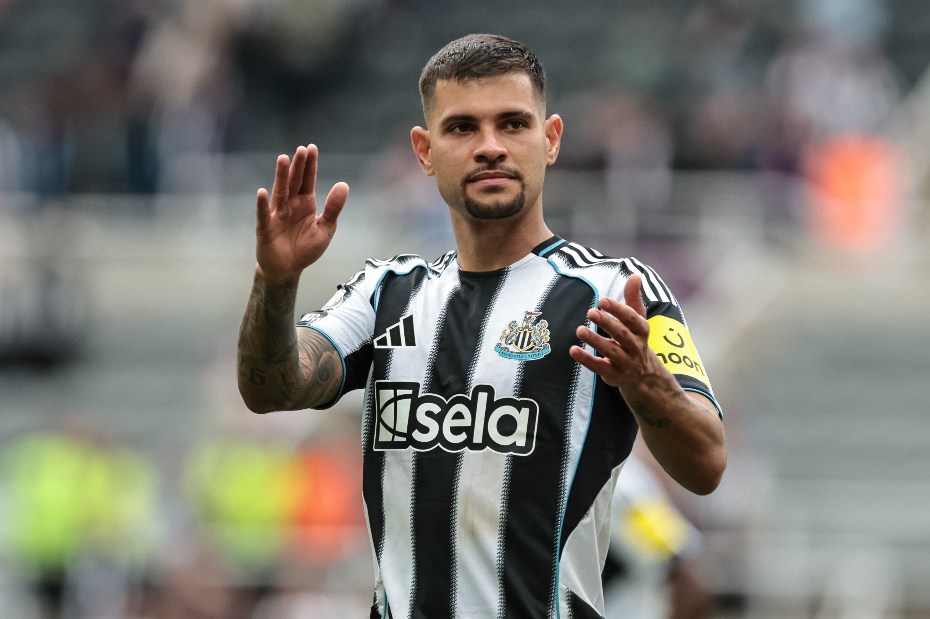 Jogador de futebol Bruno Guimarães, com barba e cabelo curto, vestindo camisa listrada preta e branca do Newcastle, com o escudo do time e o patrocínio Sela, acena com as duas mãos para o público em um estádio