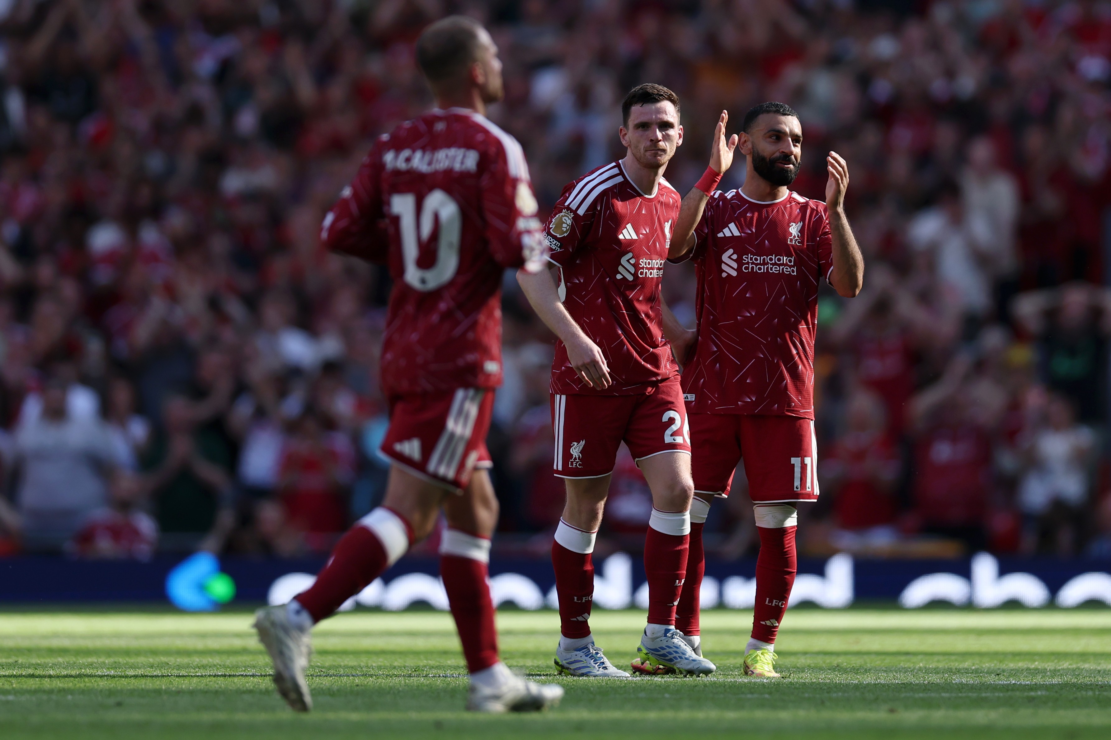 Três jogadores de futebol do Liverpool em campo, vestindo uniformes vermelhos. Um jogador de costas com a camisa 10, outro de frente com a camisa 2 e um terceiro com barba, camisa 11, aplaudindo. A torcida desfocada ao fundo