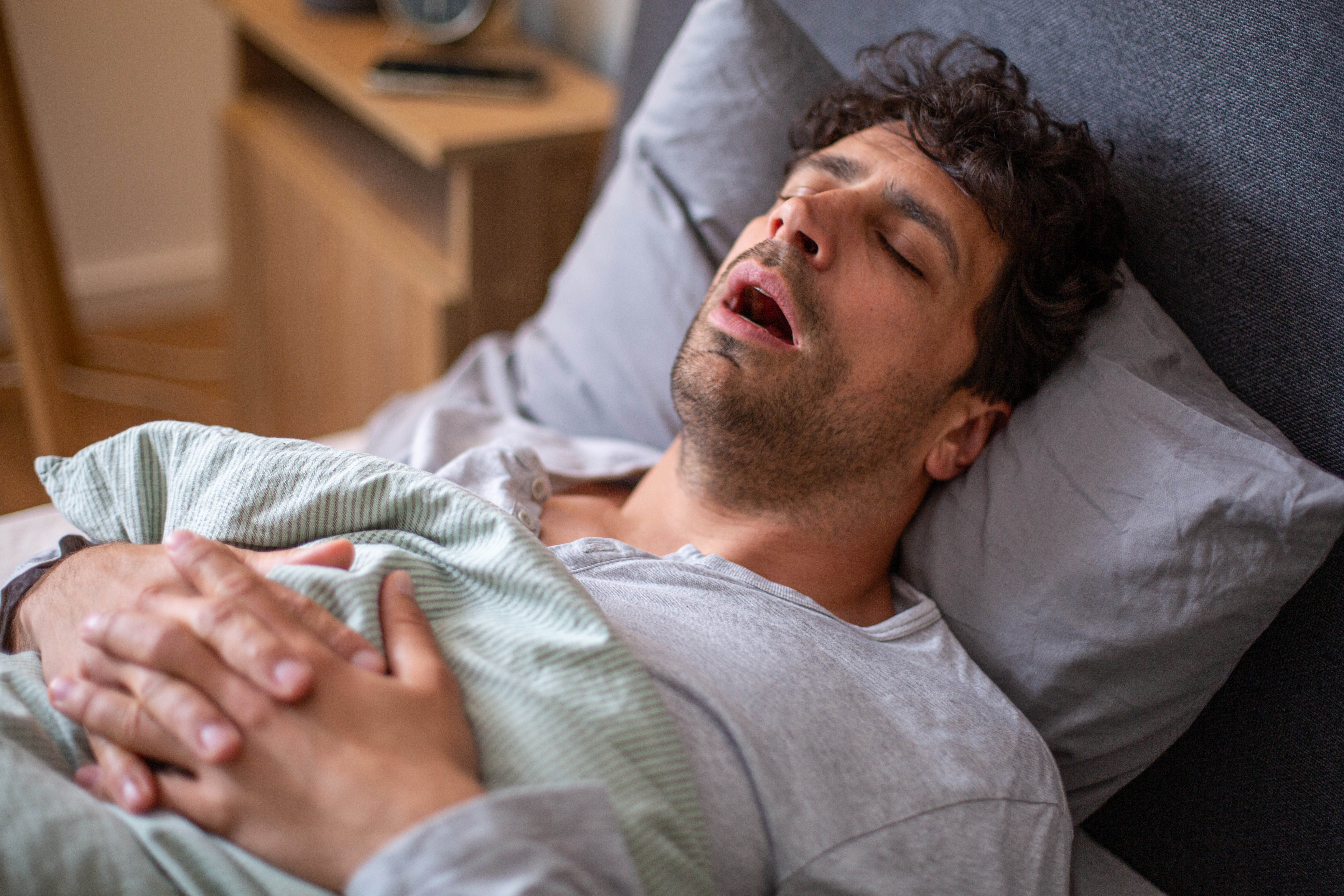 Homem deitado na cama, dormindo profundamente com a boca aberta e roncando, coberto por um lençol listrado verde e branco, com as mãos cruzadas sobre o peito