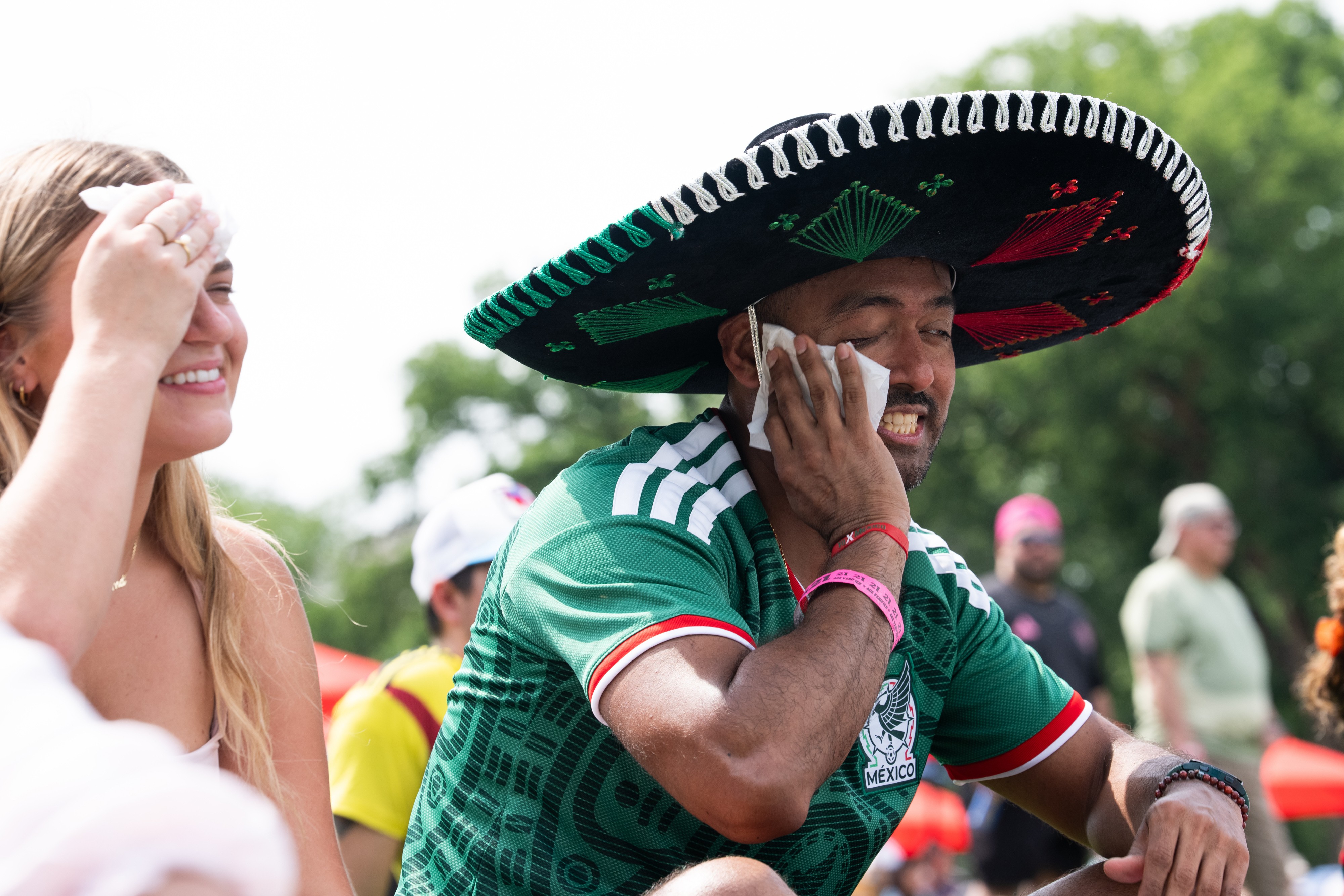 Homem de pele escura com chapéu sombrero preto e camisa verde do México, sorrindo enquanto enxuga o rosto com um lenço. Ao lado, uma mulher loira sorri, enxugando a testa com um lenço. Ambos parecem estar em um evento ao ar livre, sob o sol