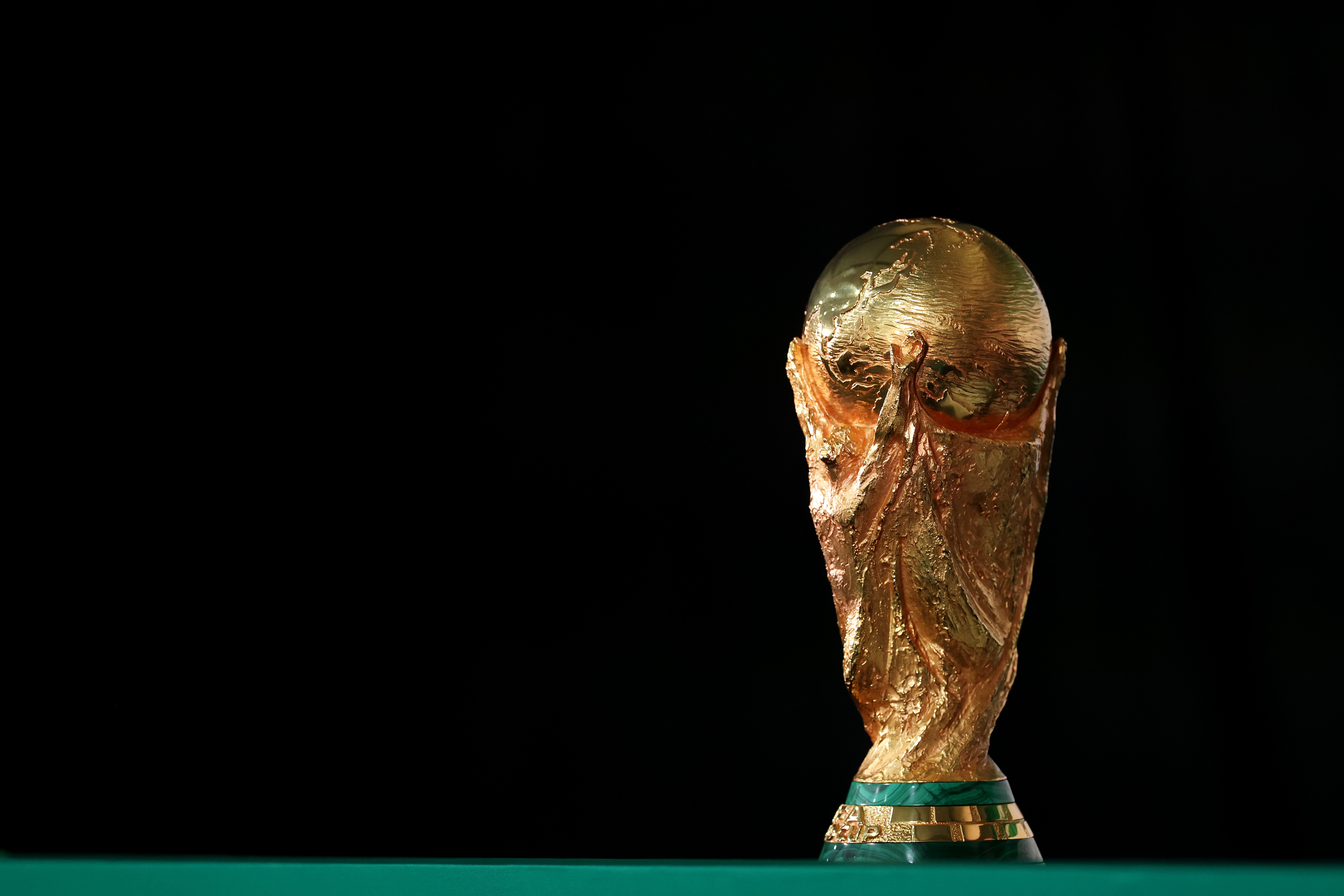 A Taça da Copa do Mundo FIFA, dourada e brilhante, em destaque sobre uma superfície verde escura, com fundo preto. O troféu tem duas figuras humanas estilizadas segurando um globo terrestre no topo