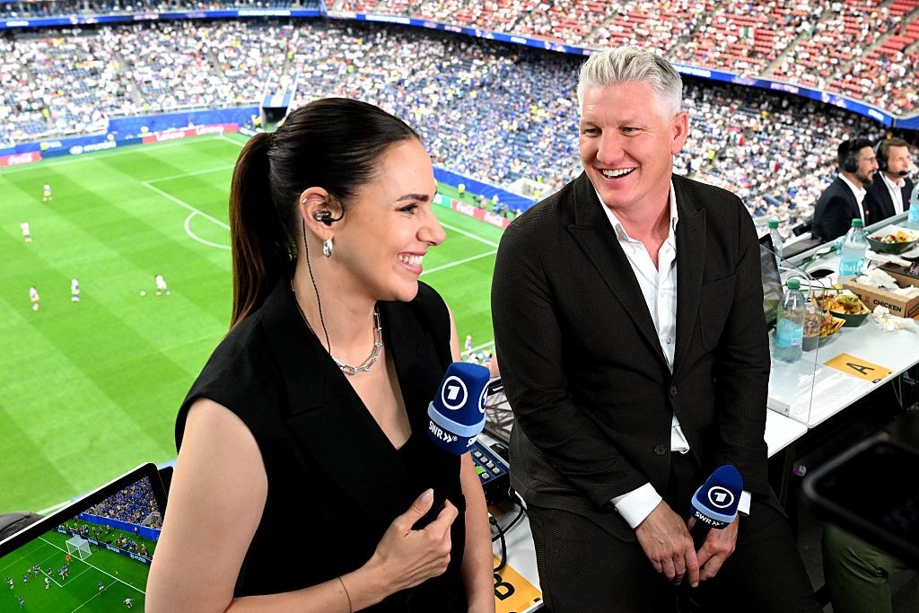 Uma mulher e um homem sorrindo em uma cabine de transmissão de futebol. Ela, à esquerda, usa blusa preta, colar prateado e fones, segurando um microfone azul. Ele, à direita, veste terno preto e camisa branca, com cabelos grisalhos. Ao fundo, o campo verde e a arquibancada lotada de um estádio