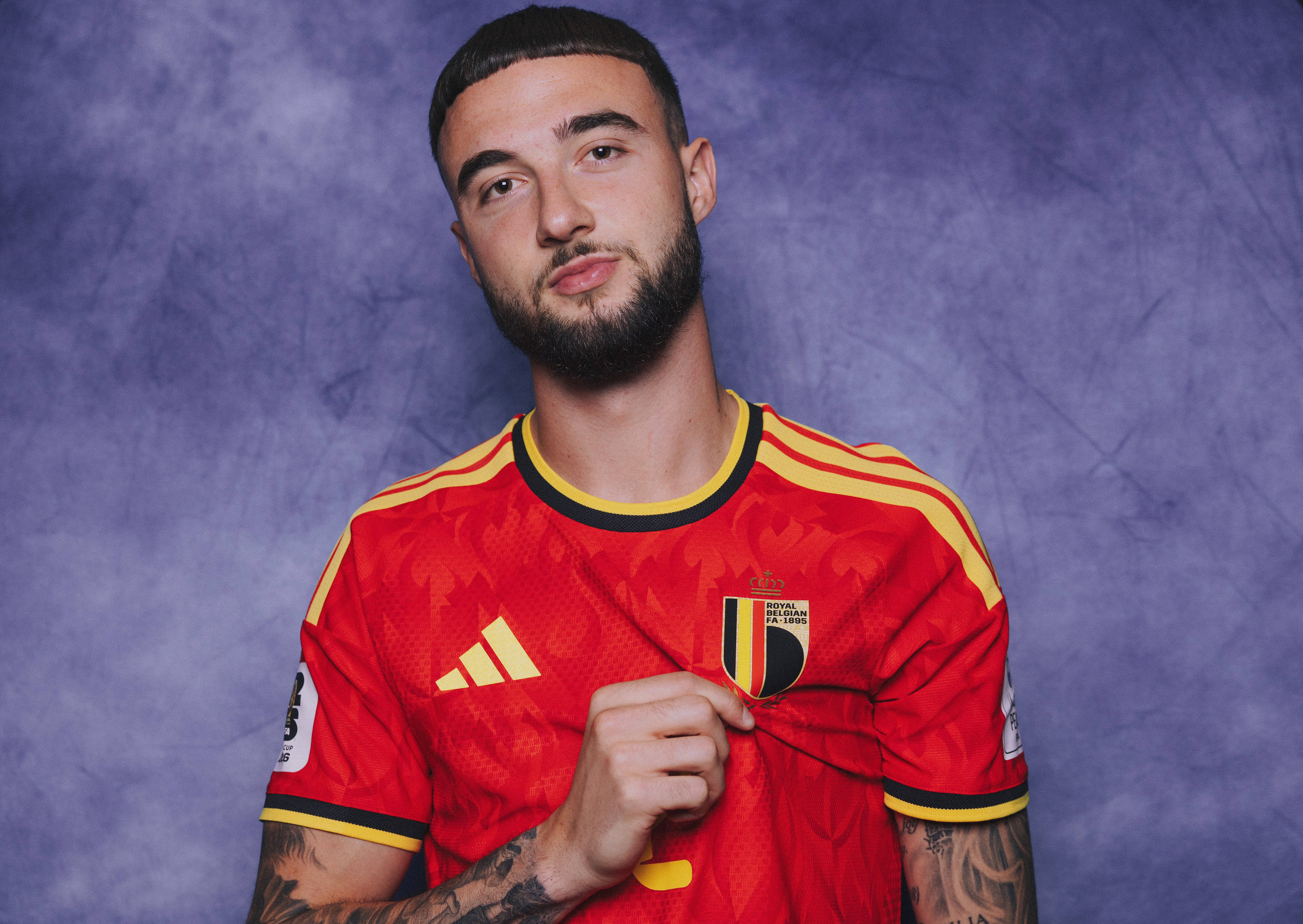 Homem jovem com barba e cabelo curto, vestindo uma camisa de futebol vermelha com detalhes amarelos e pretos, segurando o escudo da Bélgica no peito, em fundo roxo