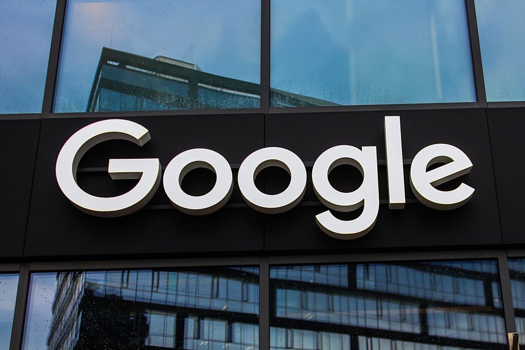 O logotipo branco da Google em relevo sobre uma fachada preta de um edifício, com reflexos de outras construções e do céu azul nas janelas de vidro acima e abaixo