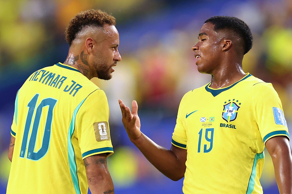 Dois jogadores de futebol brasileiros, Neymar Jr. e um colega, em campo. Neymar, &agrave; esquerda, de costas, usa camisa amarela com NEYMAR JR e 10 em azul. O outro jogador, &agrave; direita, de frente, usa camisa amarela com BRASIL e 19 em azul, gesticulando com a m&atilde;o direita aberta. Ambos est&atilde;o suados, com express&otilde;es s&eacute;rias, em um fundo desfocado de est&aacute;dio