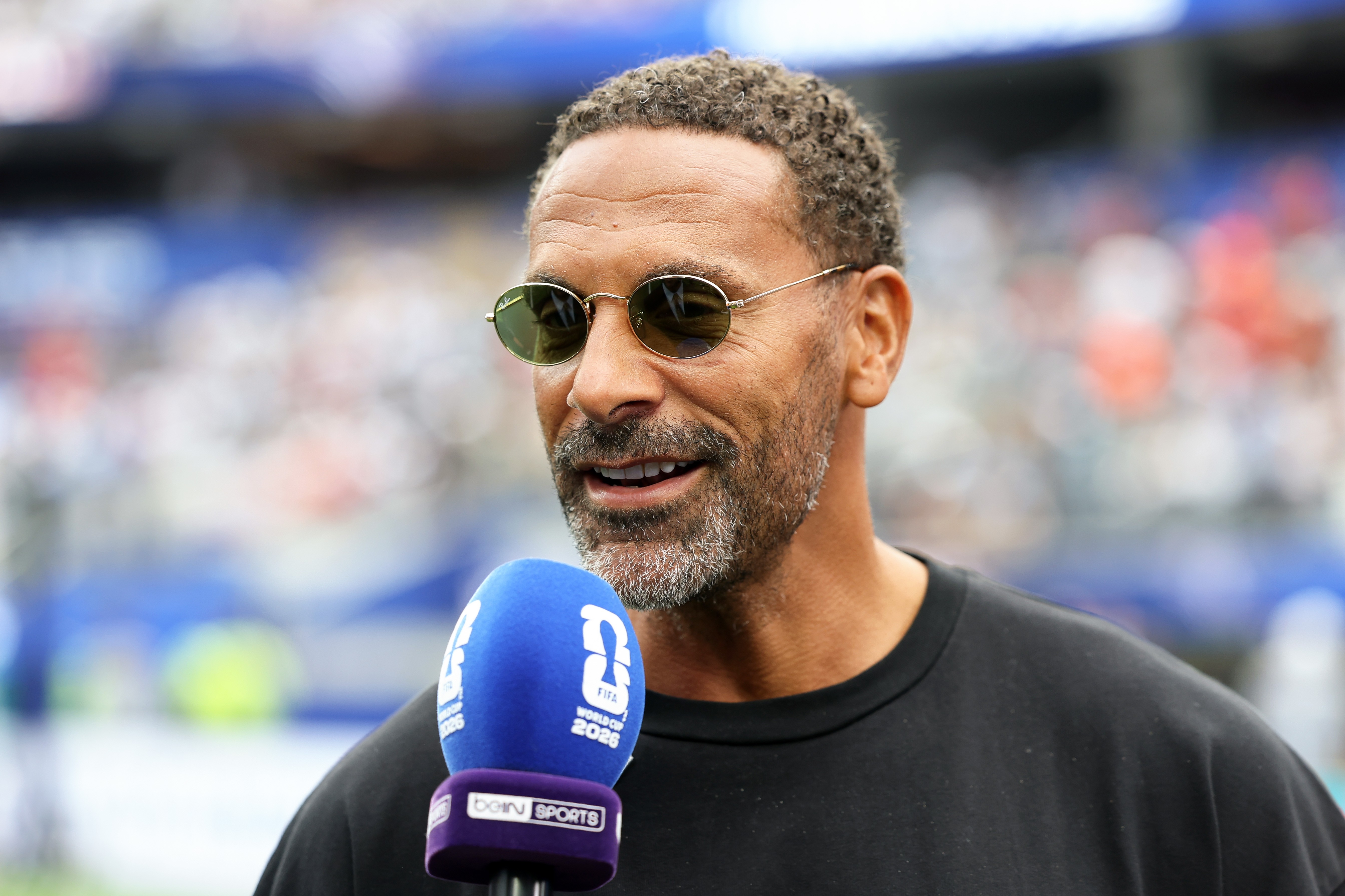 Homem negro de barba grisalha e cabelo crespo curto, usando óculos de sol redondos e camiseta preta, sorri enquanto segura um microfone azul e roxo da BeIN Sports