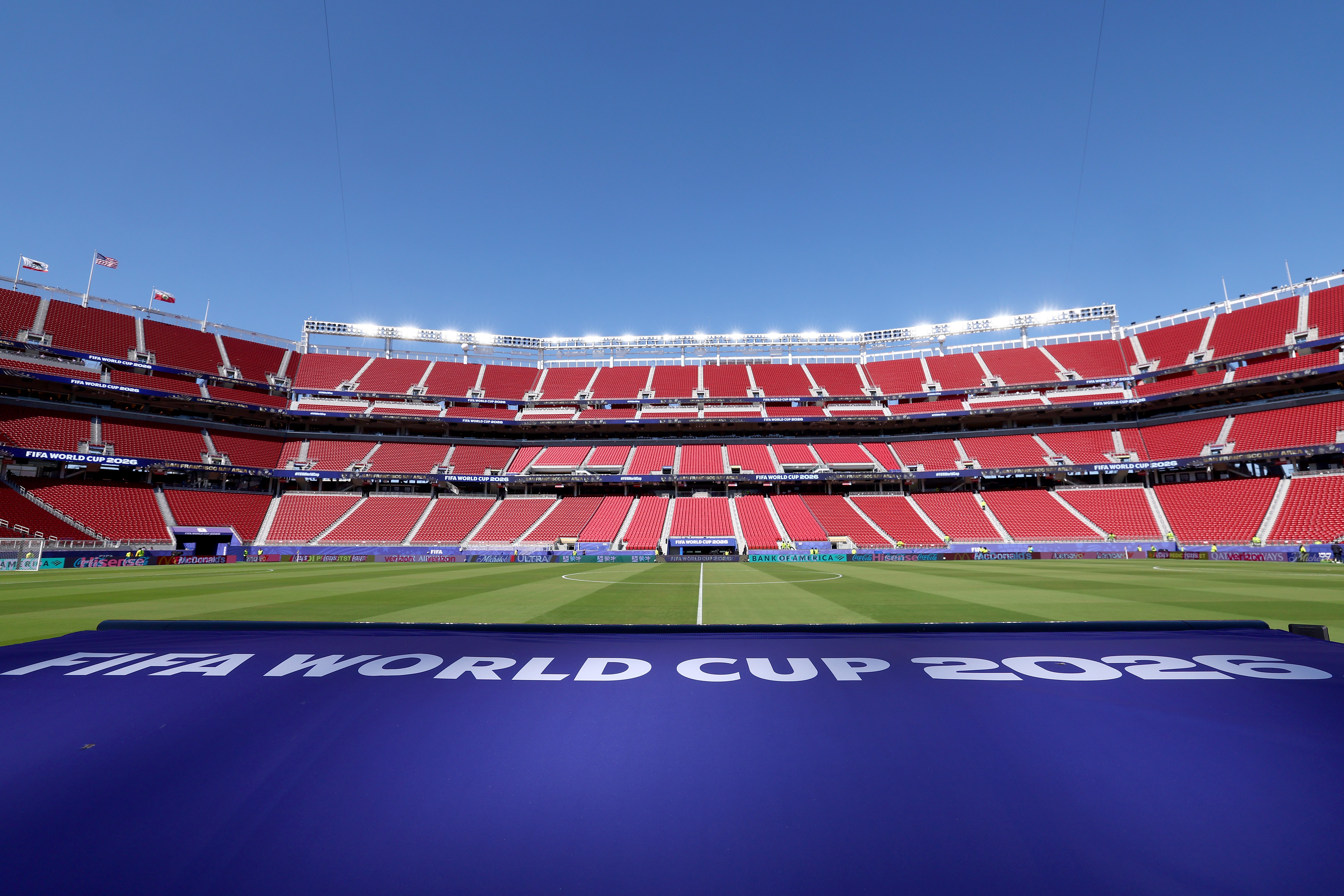 Estádio de futebol vazio com arquibancadas vermelhas em camadas, gramado verde e um banner azul no primeiro plano com FIFA WORLD CUP 2026 em branco. O céu azul claro domina o topo, e holofotes brilham no alto das arquibancadas