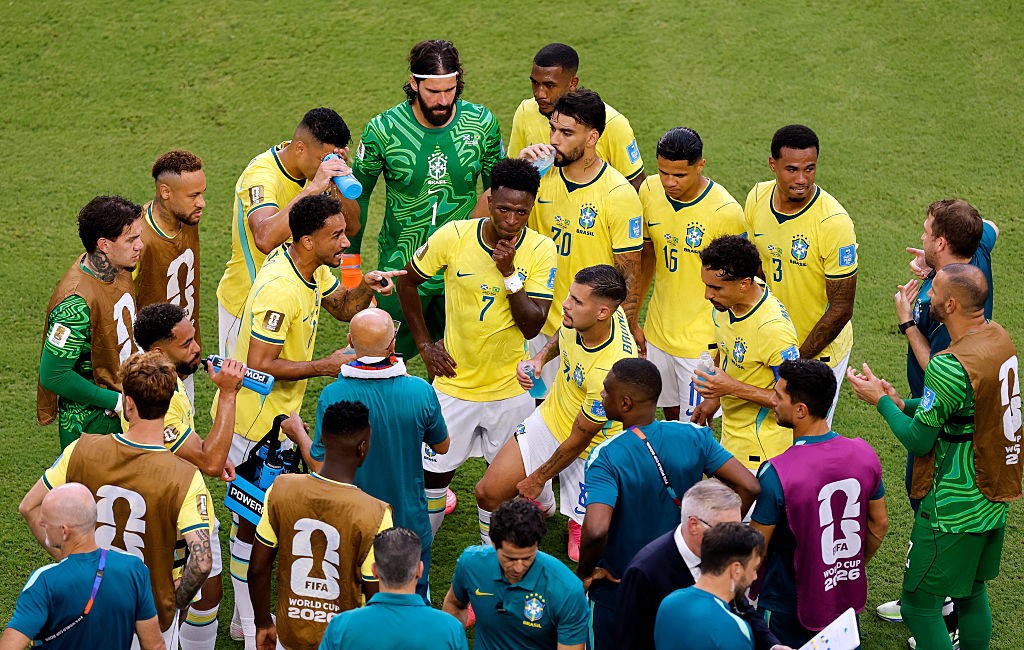 Jogadores da seleção brasileira de futebol, vestindo camisas amarelas e verdes, se reúnem em círculo no campo de grama verde, alguns bebendo água, enquanto membros da comissão técnica os observam